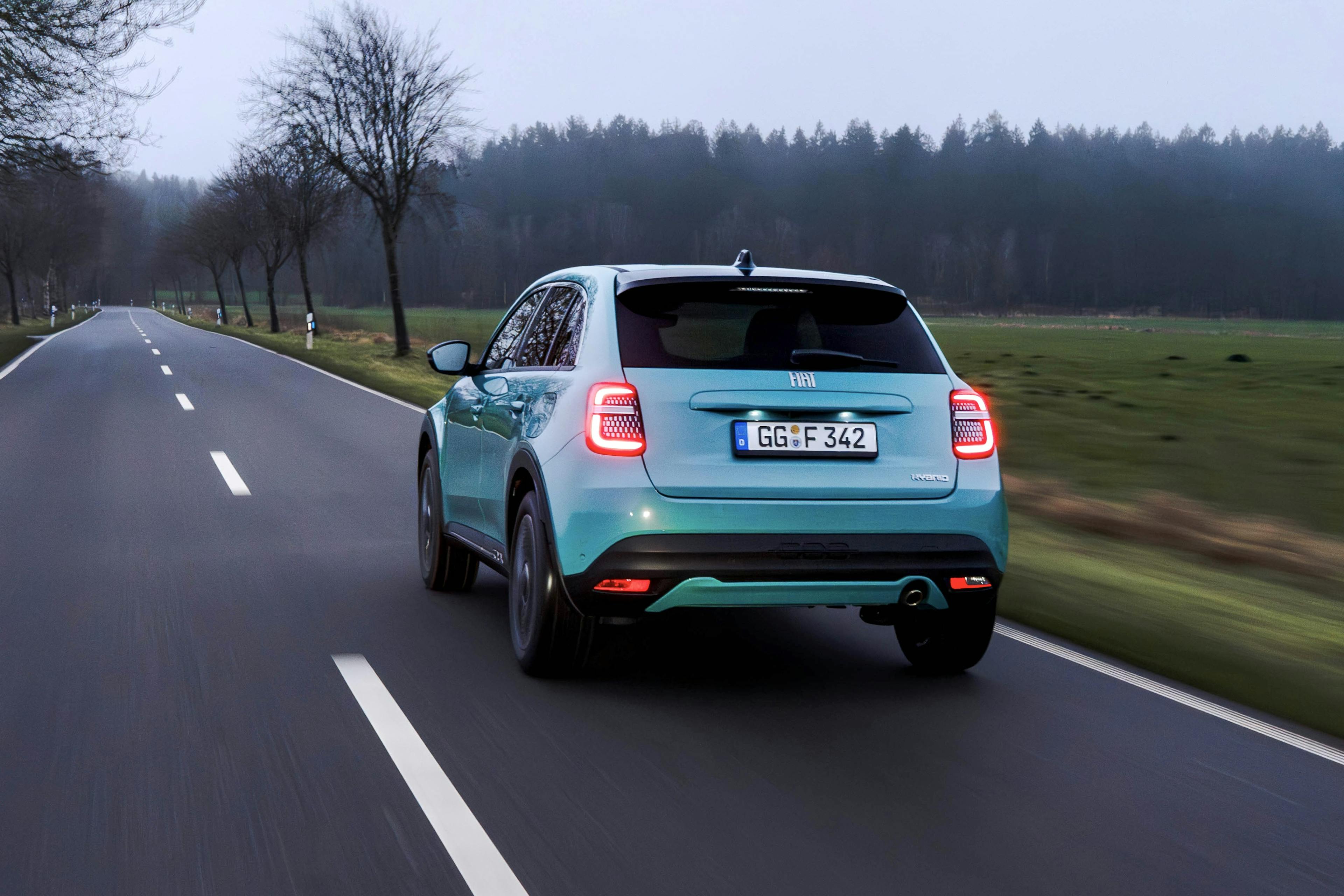 Ein blaugrüner Fiat 600 Hybrid bei Dämmerung fahrend mit eingeschalteten Rückleuchten auf einer Landstraße mit laublosen Bäumen im Hintergrund.