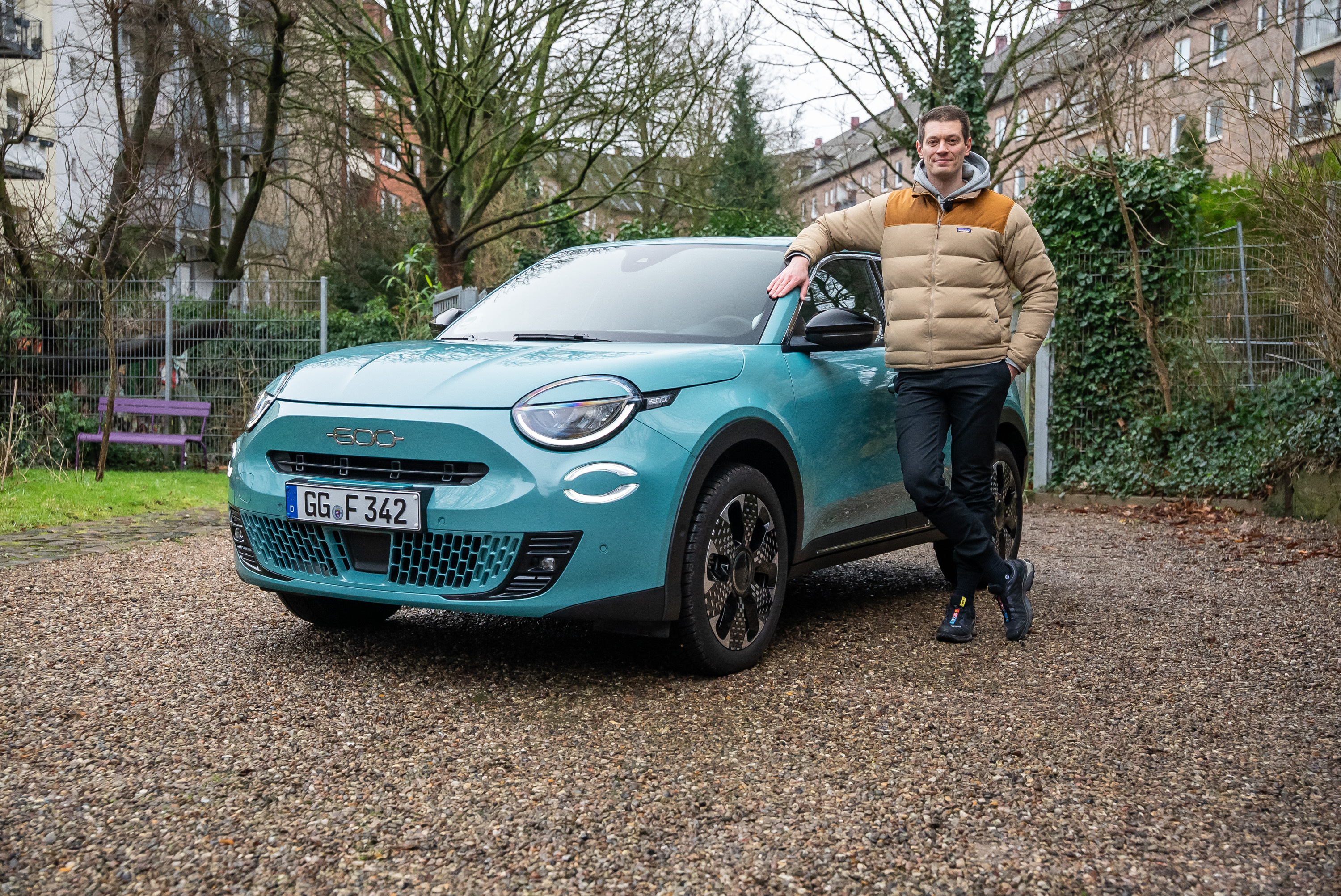 Ein Mann steht lächelnd neben einem türkisfarbenen Fiat 500 mit deutschem Kennzeichen, das auf einem Kiesplatz vor einer Wohnhauskulisse geparkt ist.