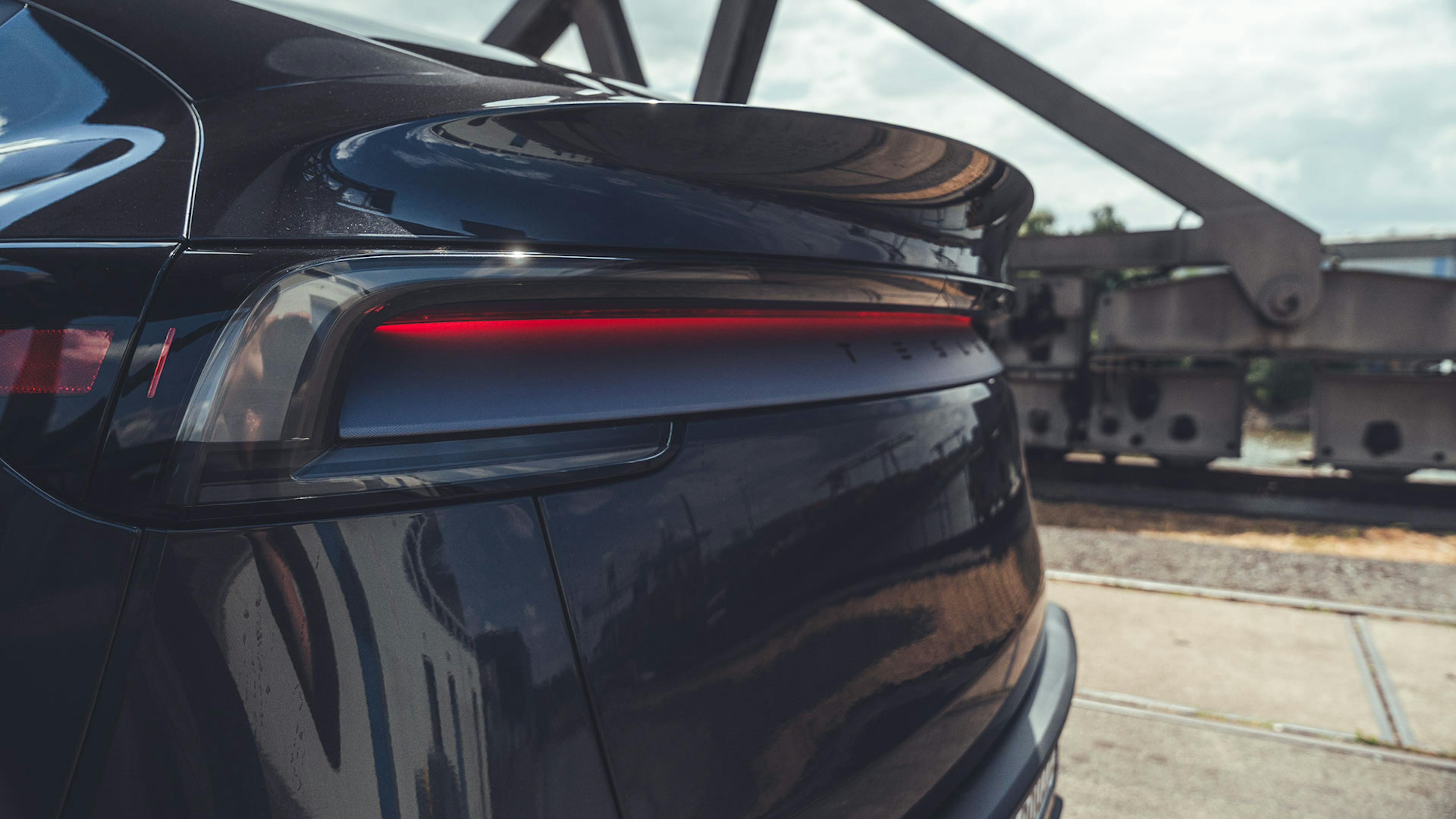 Ein dunkelgraues Tesla Model Y parkt im Sonnenschein in einem Hafengebiet. Das indirekte rote Licht der Rückleuchte ist eingeschaltet. 