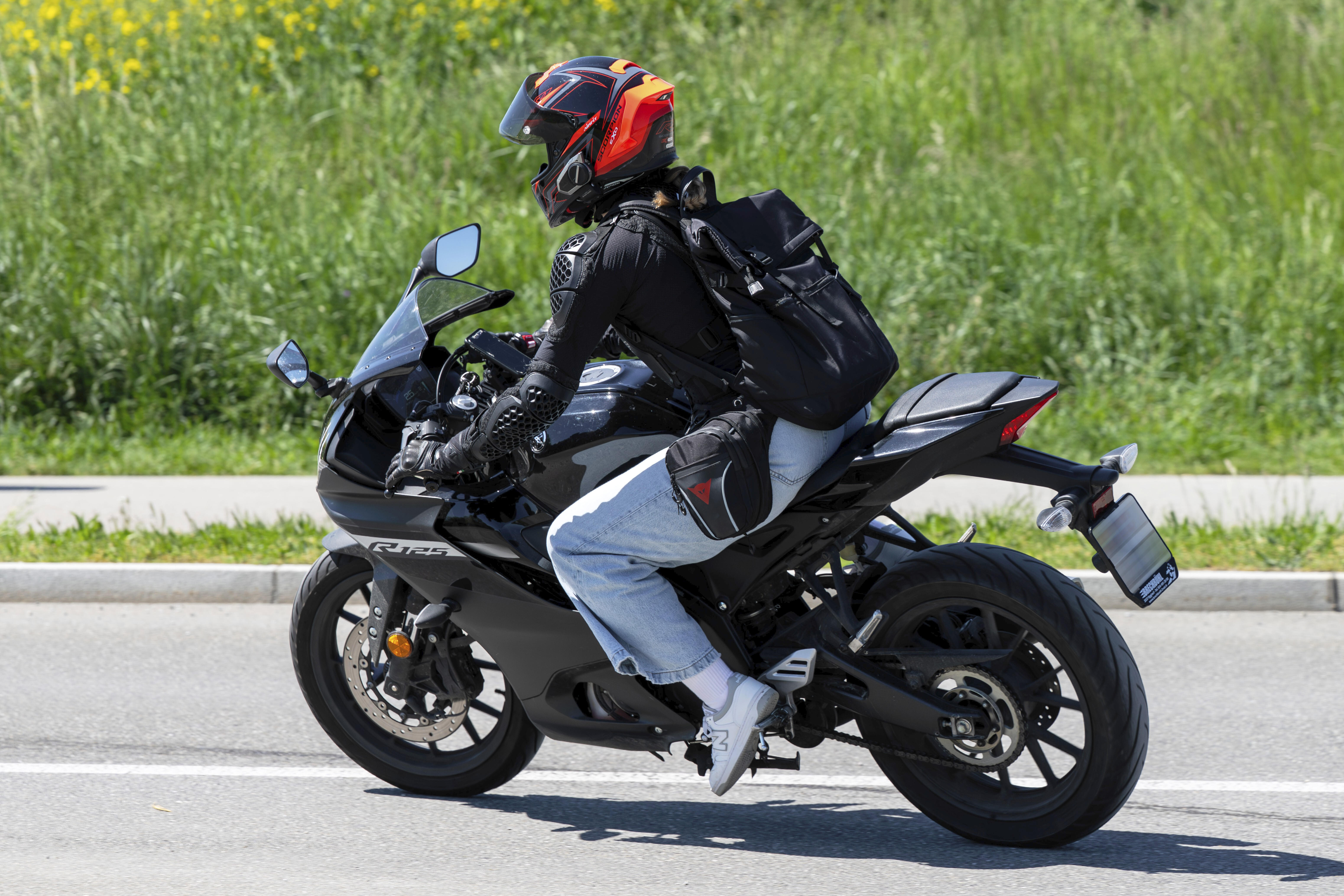 Eine jüngere Person fährt auf einem Motorrad. Sie trägt eine Jeans, eine Motorradjacke und einen schwarz-orangefarbenen Helm.