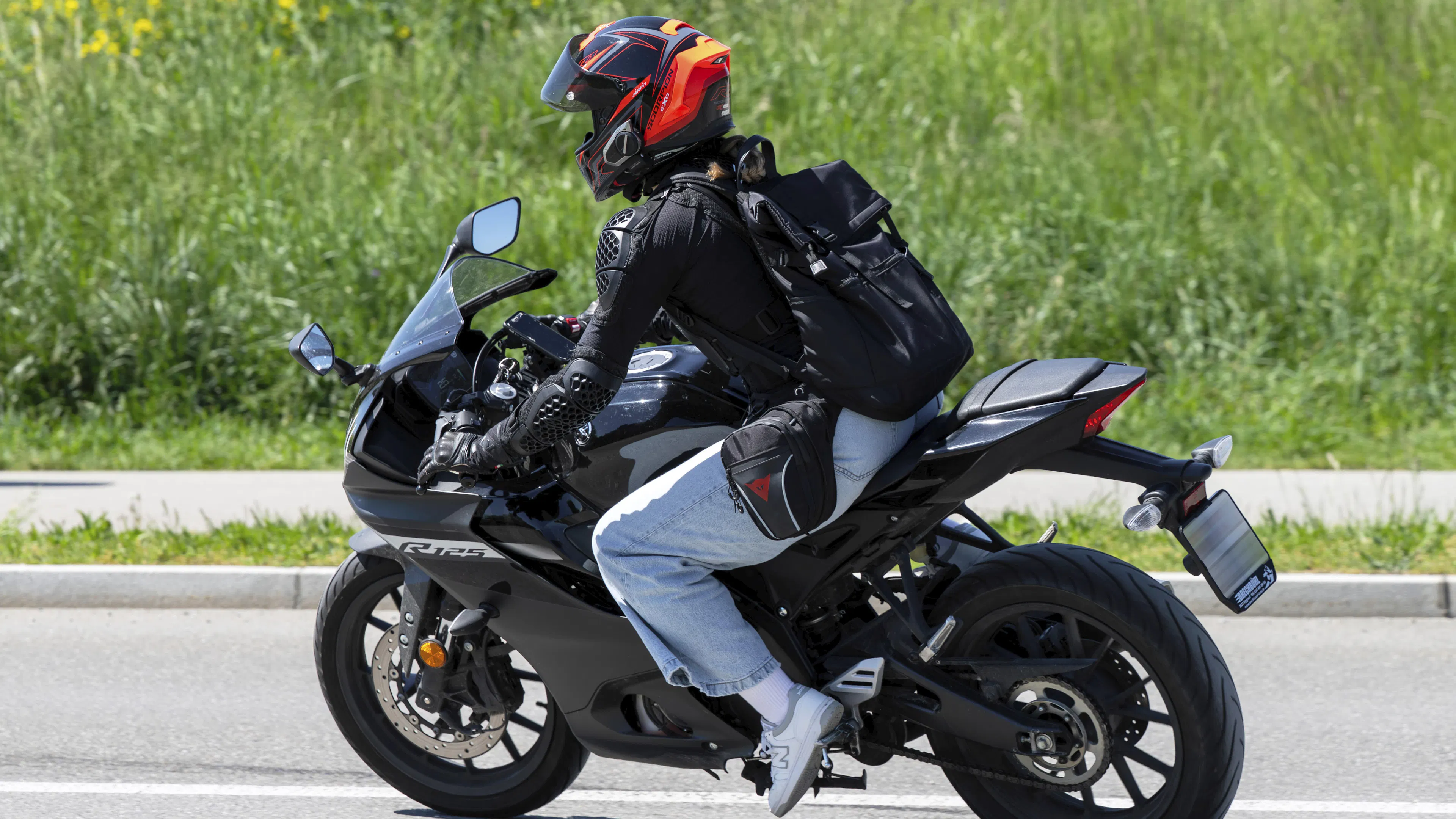 Eine jüngere Person fährt auf einem Motorrad. Sie trägt eine Jeans, eine Motorradjacke und einen schwarz-orangefarbenen Helm.