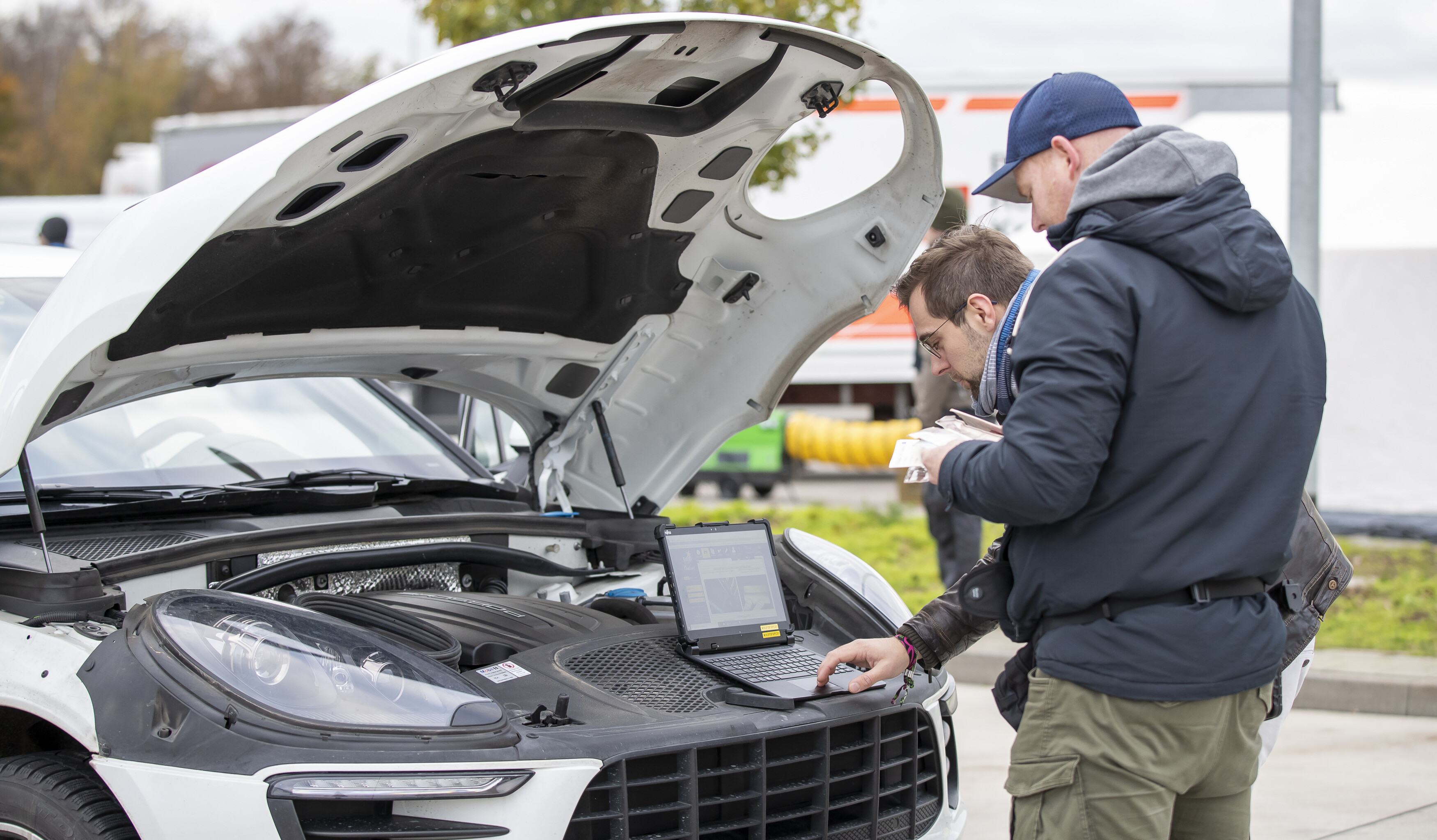 Zwei Männer stehen an einem Wagen mit geöffneter Motorhaube. Sie fragen eine Fahrzeug-Identifizierungsnummer mit dem Computer ab. 
