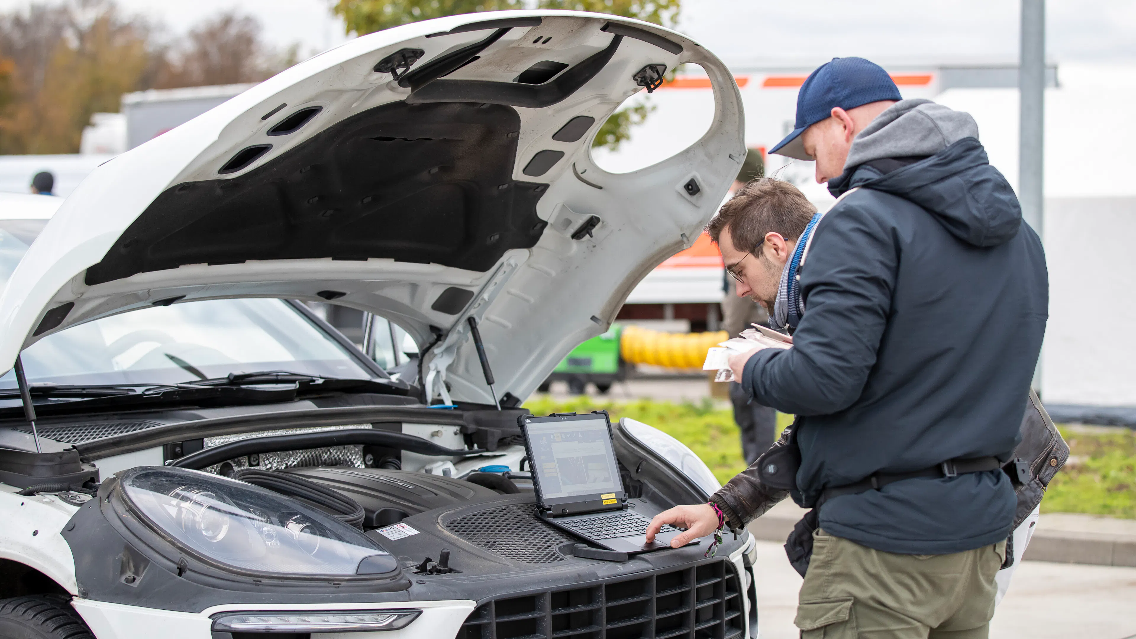 Zwei Männer stehen an einem Wagen mit geöffneter Motorhaube. Sie fragen eine Fahrzeug-Identifizierungsnummer mit dem Computer ab.