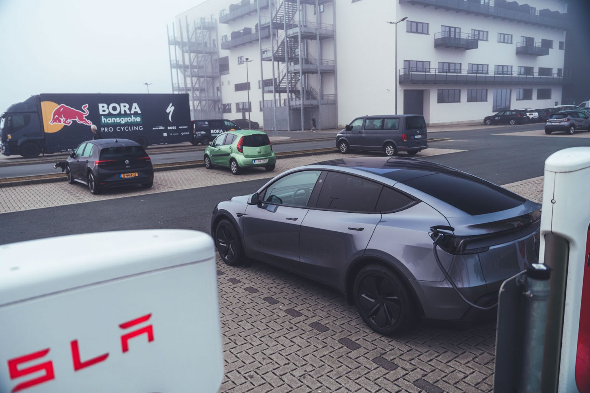 Ein dunkelgraues Tesla Model Y parkt im im Nebel an einer Ladesäule. Im Hintergrund stehen LKW und weitere Autos. 