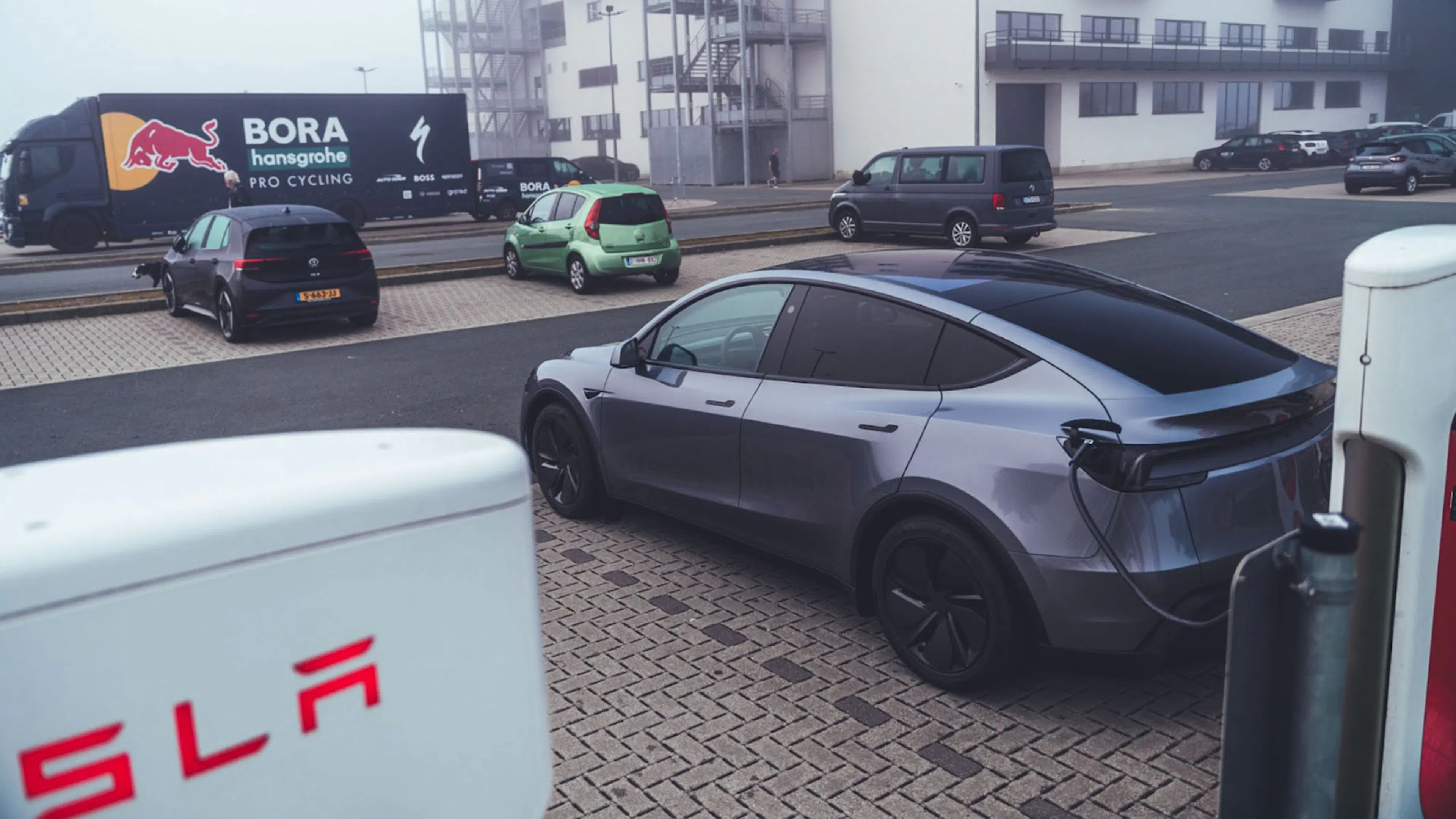Ein dunkelgraues Tesla Model Y parkt im im Nebel an einer Ladesäule. Im Hintergrund stehen LKW und weitere Autos.