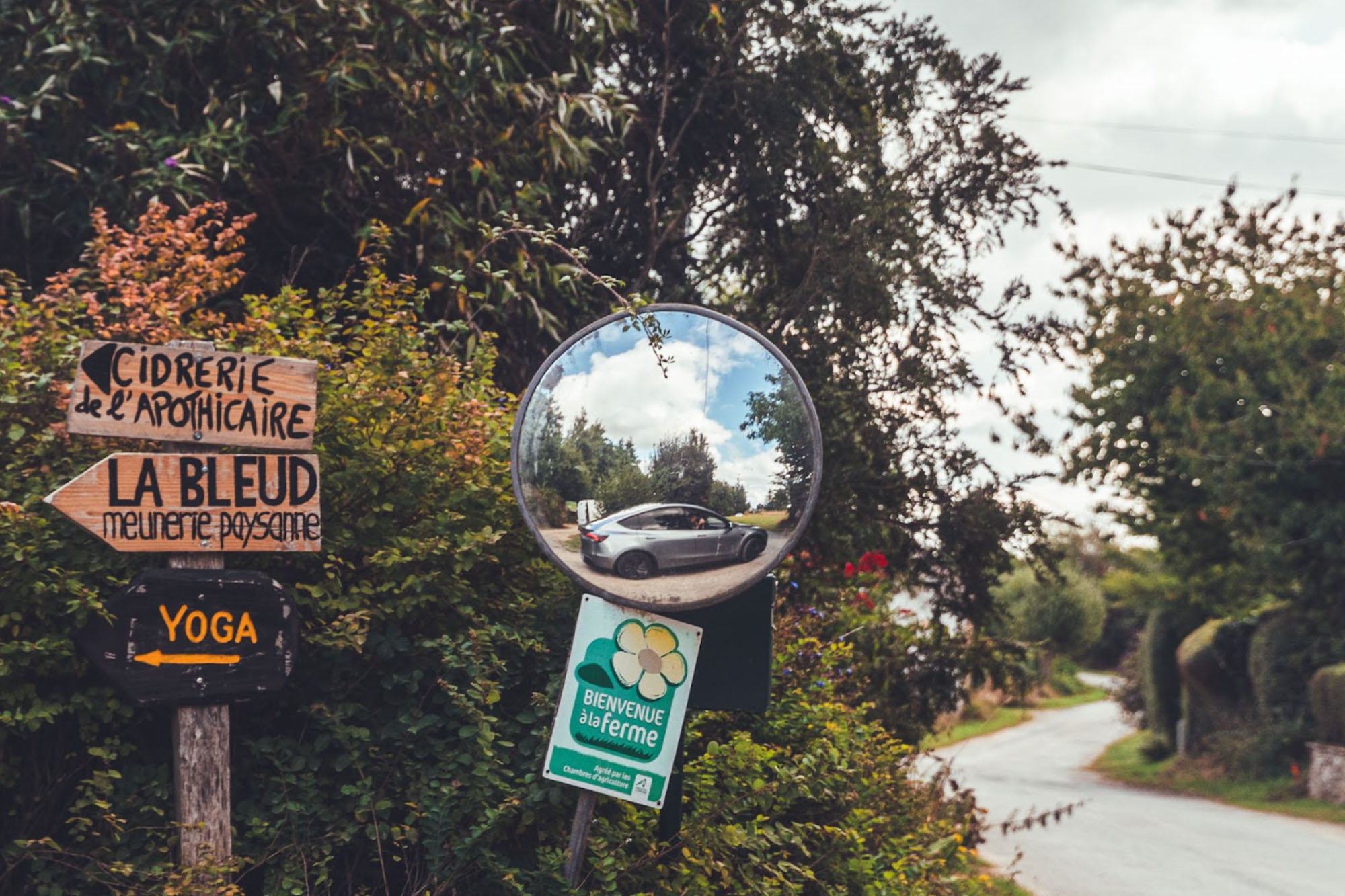  Wegweiser aus Holz zeigen nach links während in einem Spiegel ein Tesla Model Y auf einer Kreuzung abbiegen will.