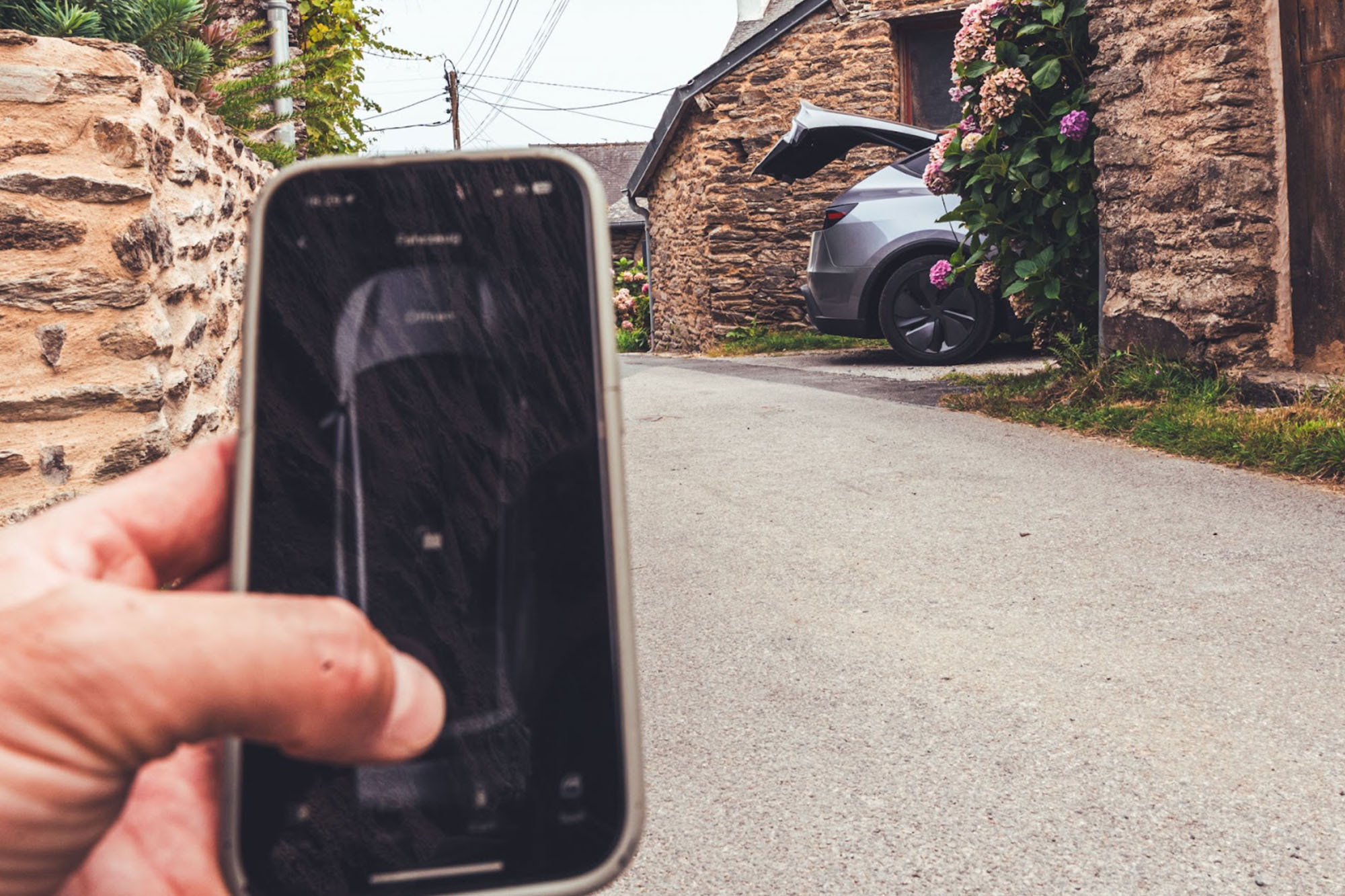 Im Vordergrund hält eine Hand ein Smartphone während sich im Hintergrund die Kofferraumklappe eines Tesla Model Y öffnet