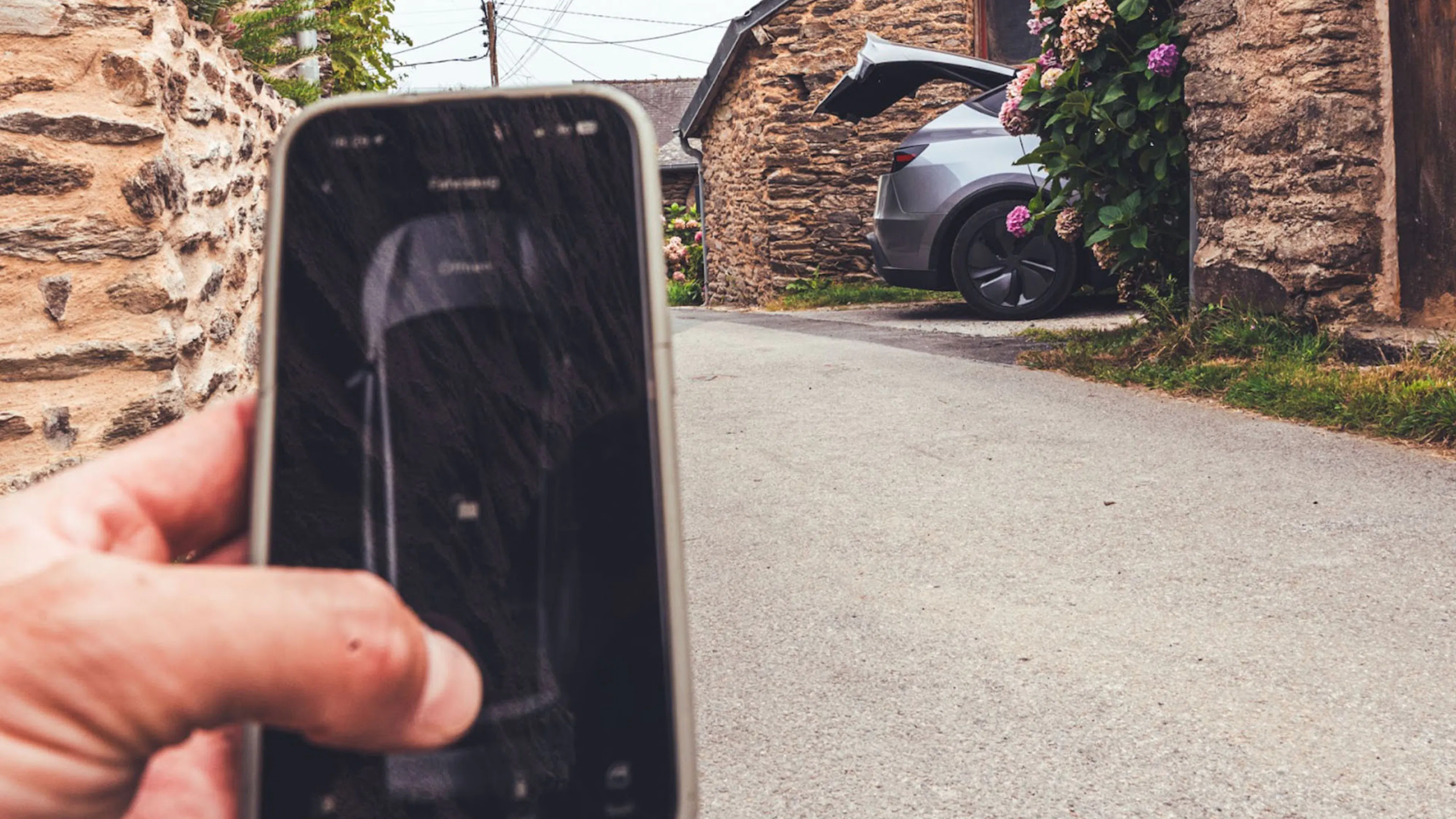 Im Vordergrund hält eine Hand ein Smartphone während sich im Hintergrund die Kofferraumklappe eines Tesla Model Y öffnet