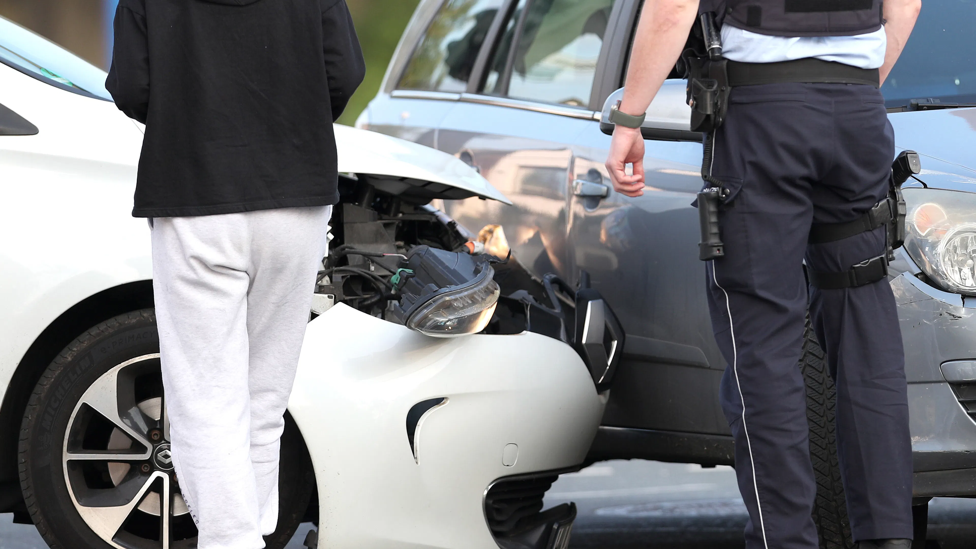 Ein beschädigtes weißes Auto steht nach einem Unfall mit einem silbernen Auto auf der Straße. Ein Polizist und eine Frau stehen davor.