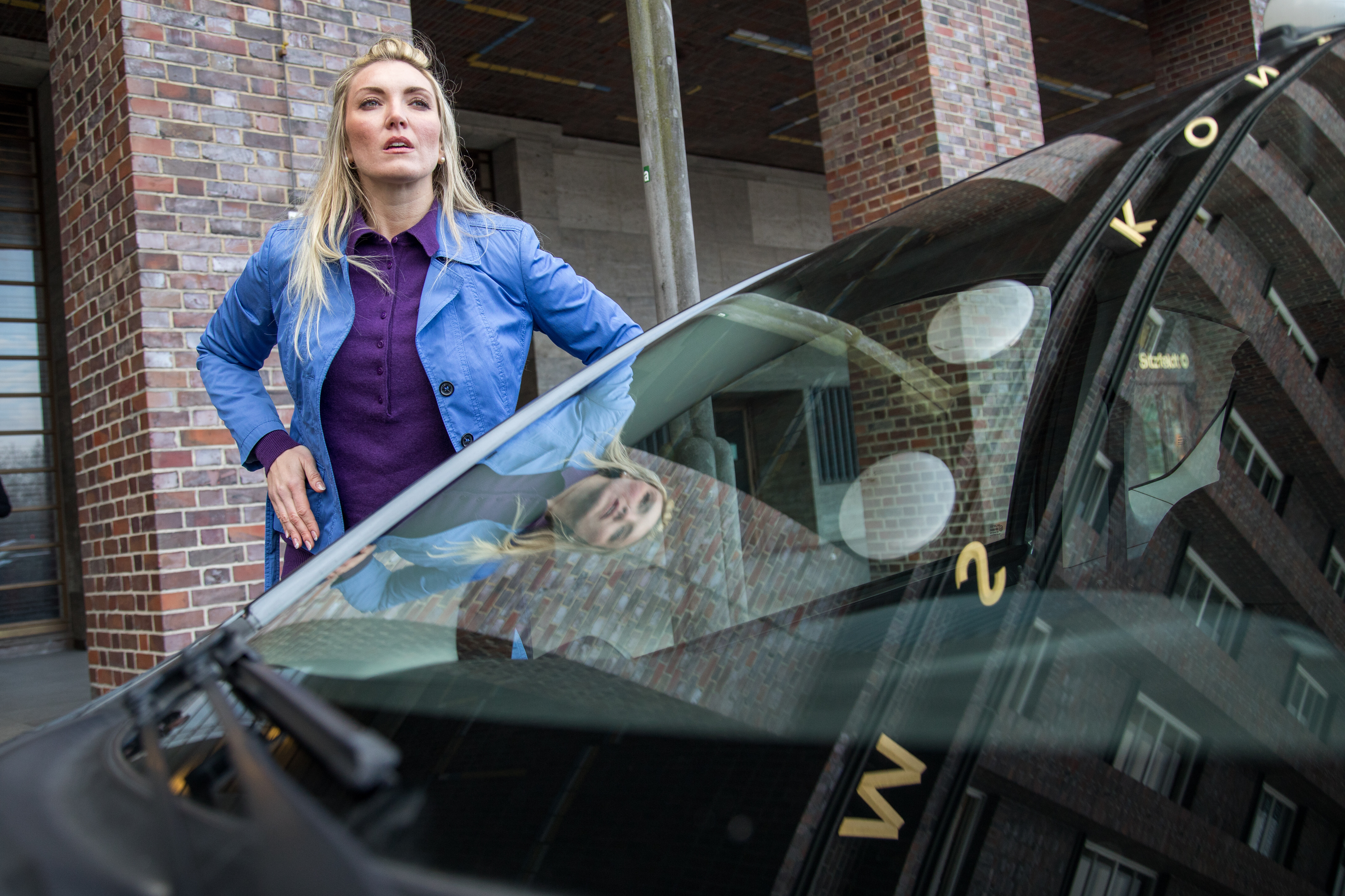 Eine Frau mit eine blauen Jacke und lilafarbenen Bluse steht vor einem dunklen Auto und sucht ihren Autoschlüssel.