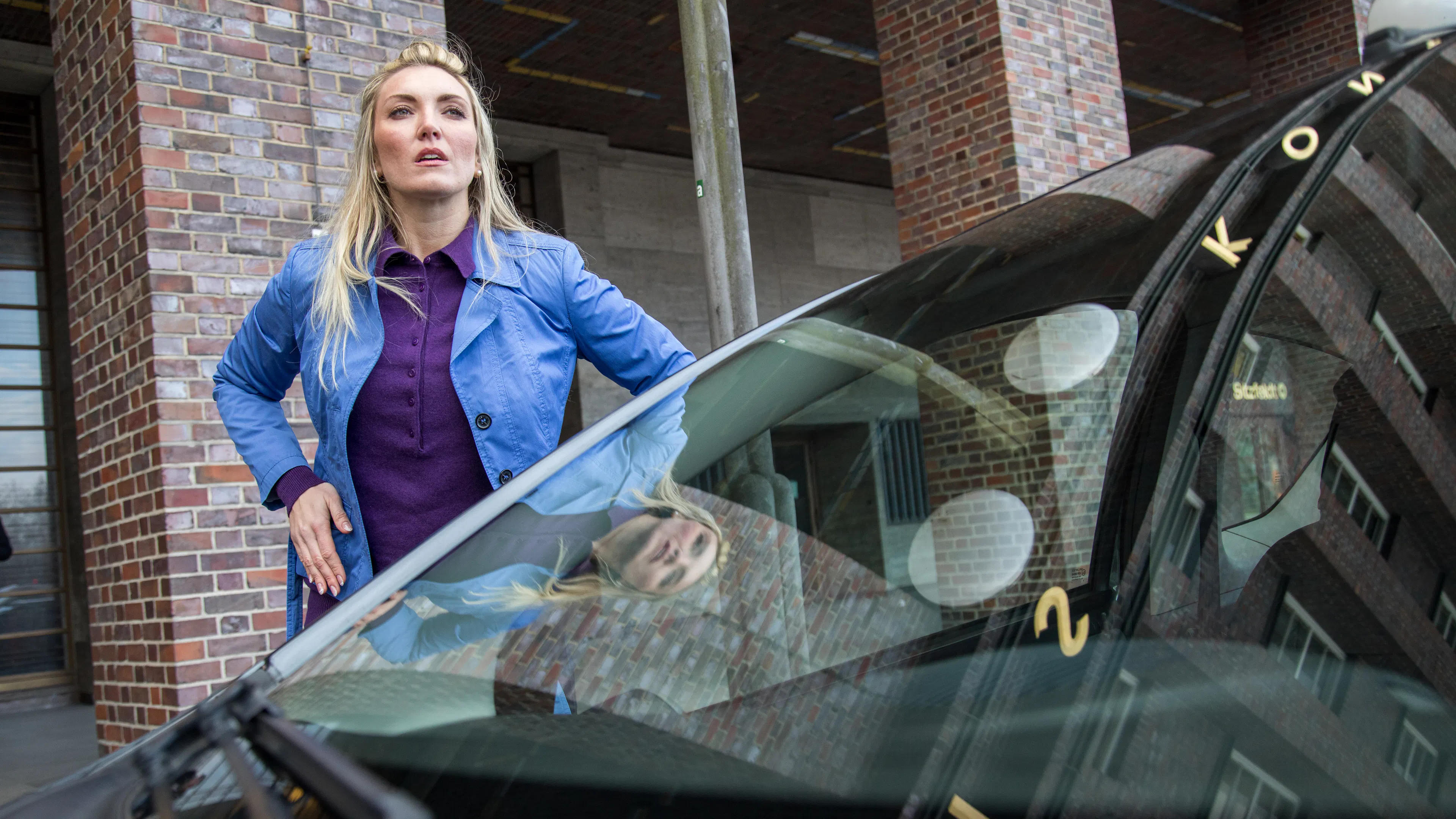 Eine Frau mit eine blauen Jacke und lilafarbenen Bluse steht vor einem dunklen Auto und sucht ihren Autoschlüssel.