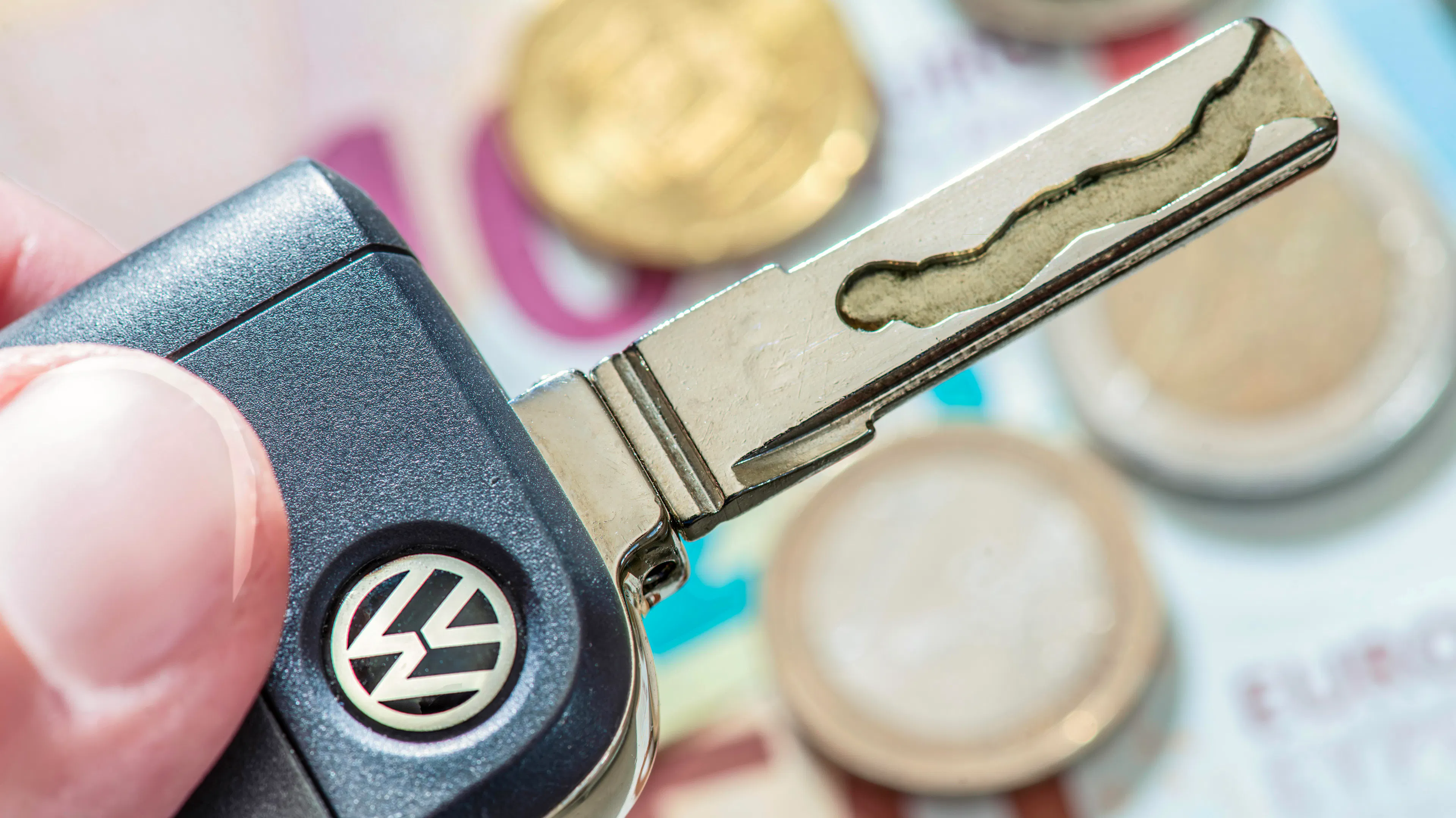 Jemand hält einen Autoschlüssel von VW in der Hand. Im Hintergrund sieht man Geldscheine und Münzen.