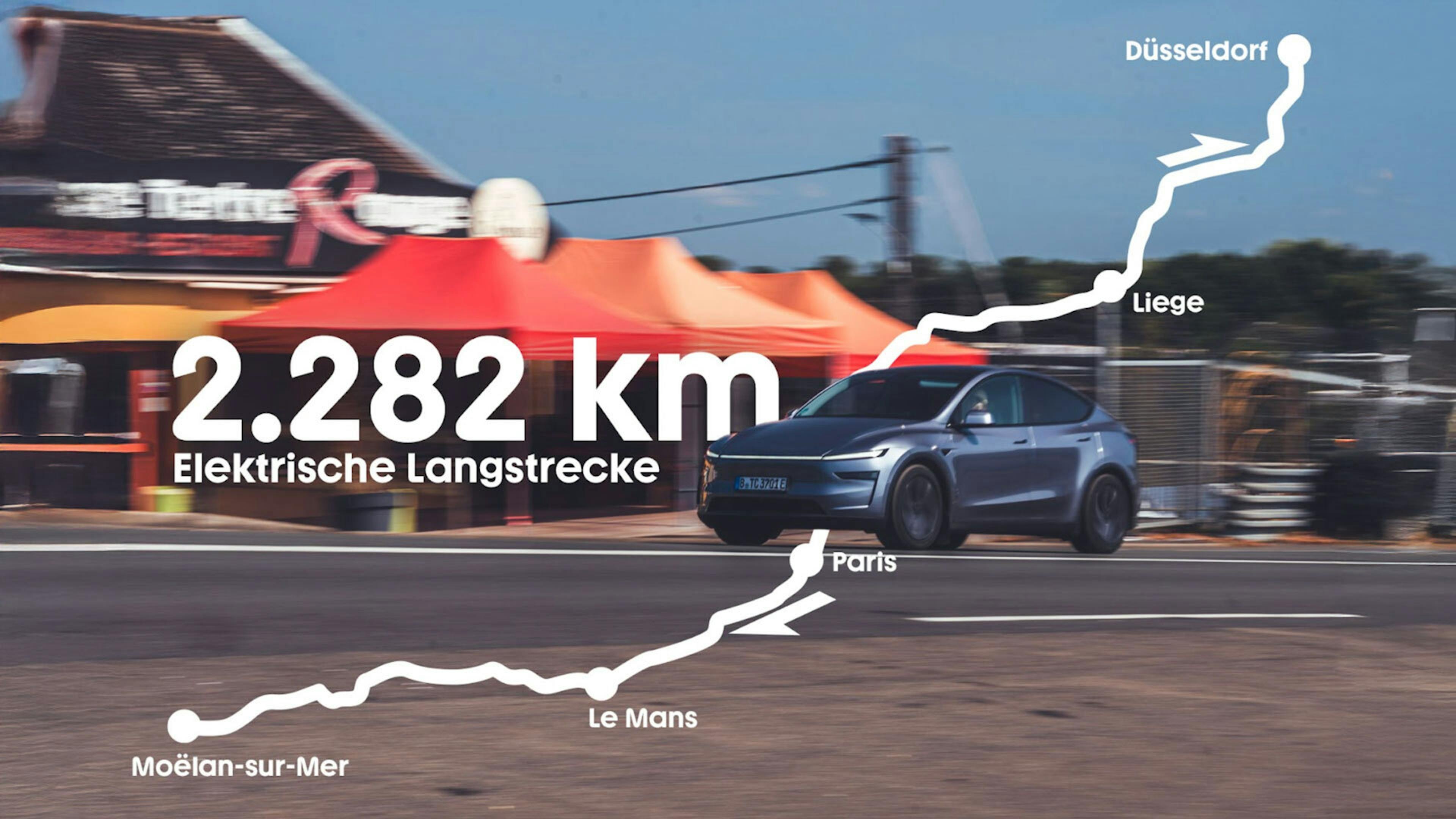 Ein dunkelgraues Tesla Model Y fährt auf einer Landstraße an einem Restaurant in Le Mans vorbei. Eine weiße Grafik beschreibt die 2.282 km weite Strecke des Hin- und Rückweges.