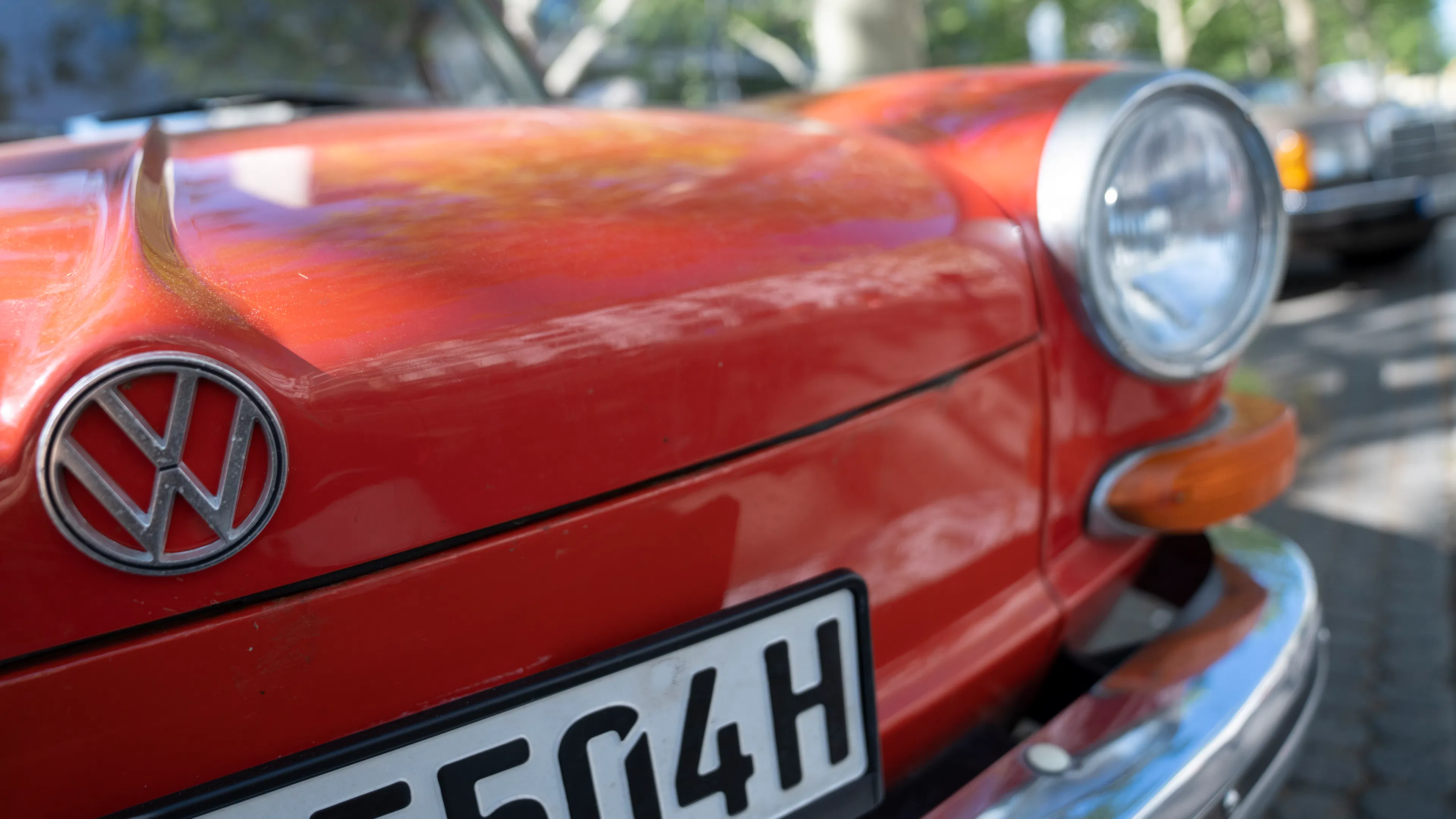 Ein roter VW-Oldtimer mit H-Kennzeichen, geparkt in einer schattigen Umgebung mit Bäumen im Hintergrund.