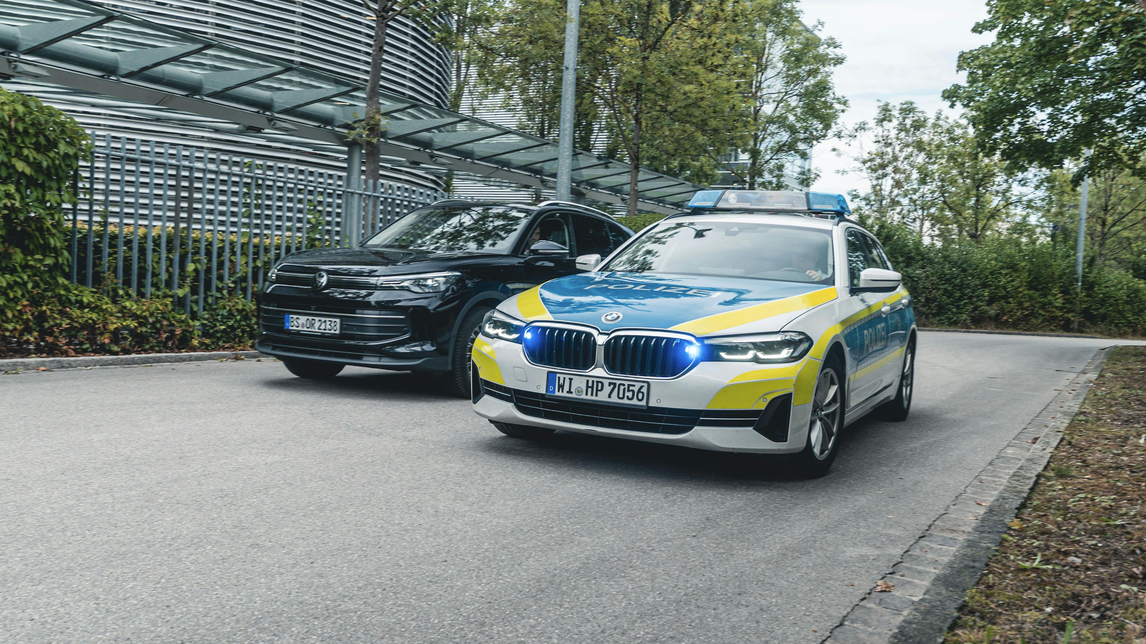 Ein Polizeiauto überholt einen dunklen VW, um das C-ITS zu demonstrieren: "Bildung einer Rettungsgasse!