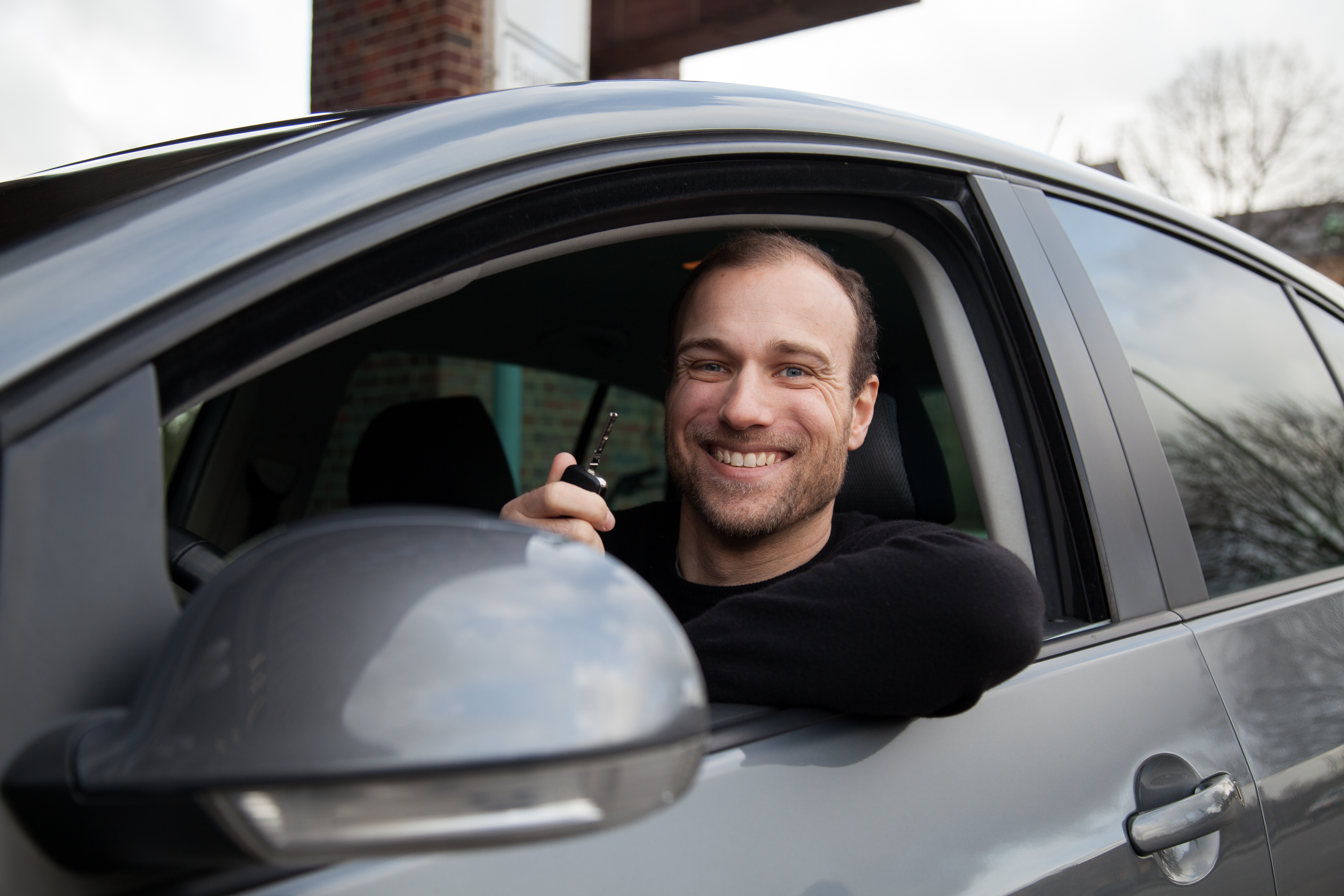 Ein junger Mann lächelt aus dem Auto heraus und hält den Autoschlüssel in der Hand.