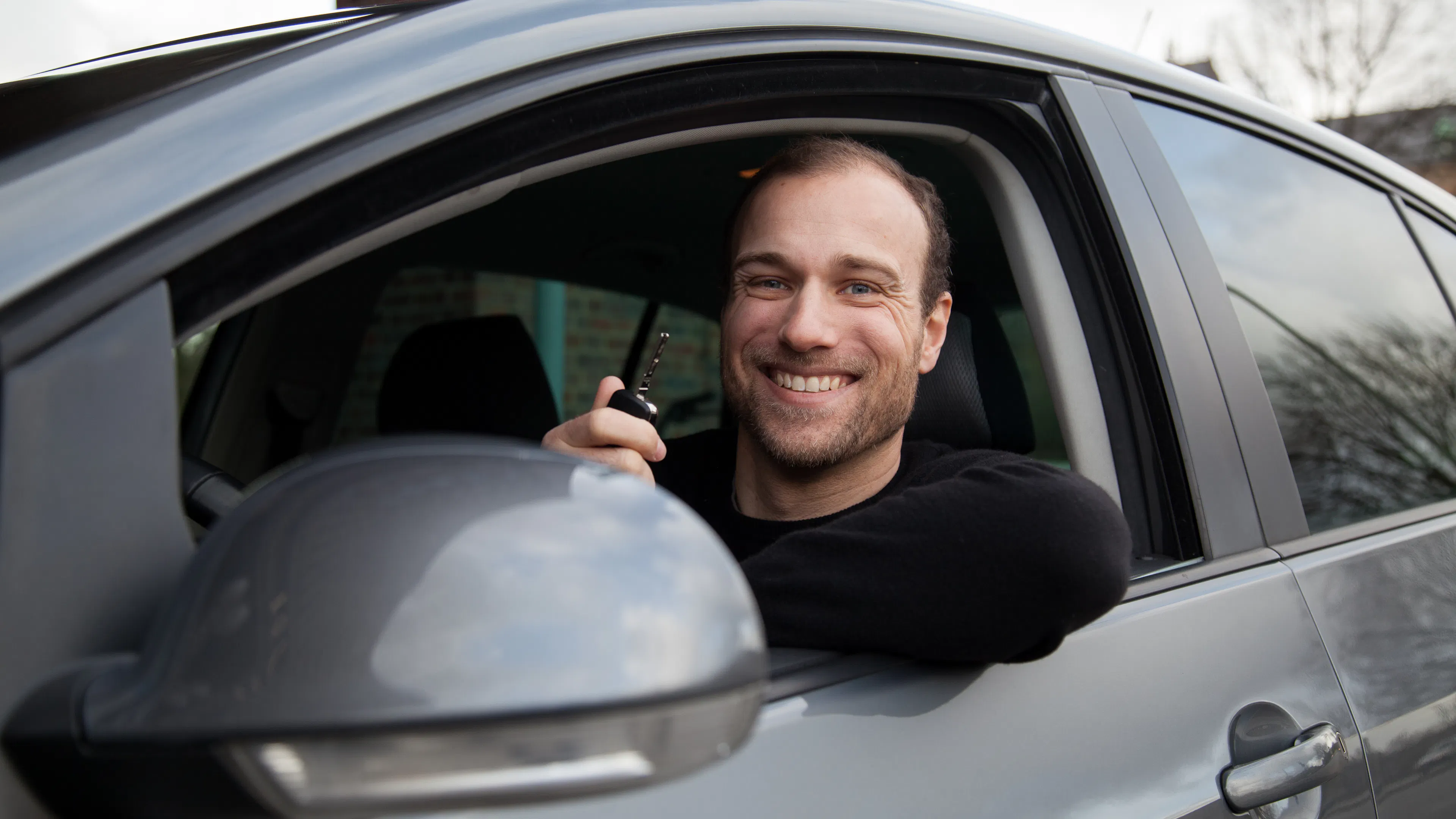 Ein junger Mann lächelt aus dem Auto heraus und hält den Autoschlüssel in der Hand.