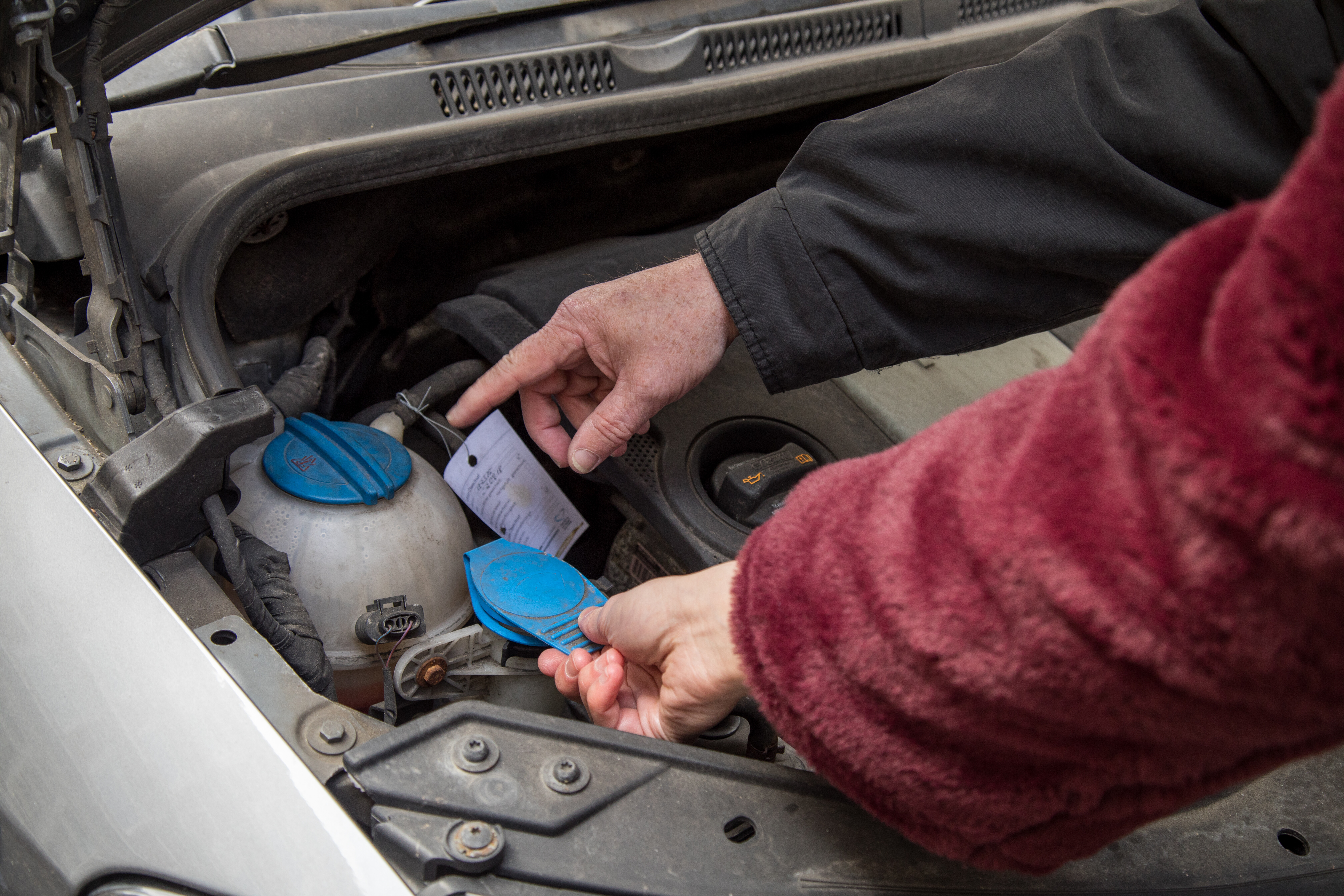 Zwei Personen sehen den Motorraum eines Autos an. Die eine Person zeigt auf den Kühlmittelbehälter, die andere fasst den Deckel des Scheibenwischwasserbehälters an.