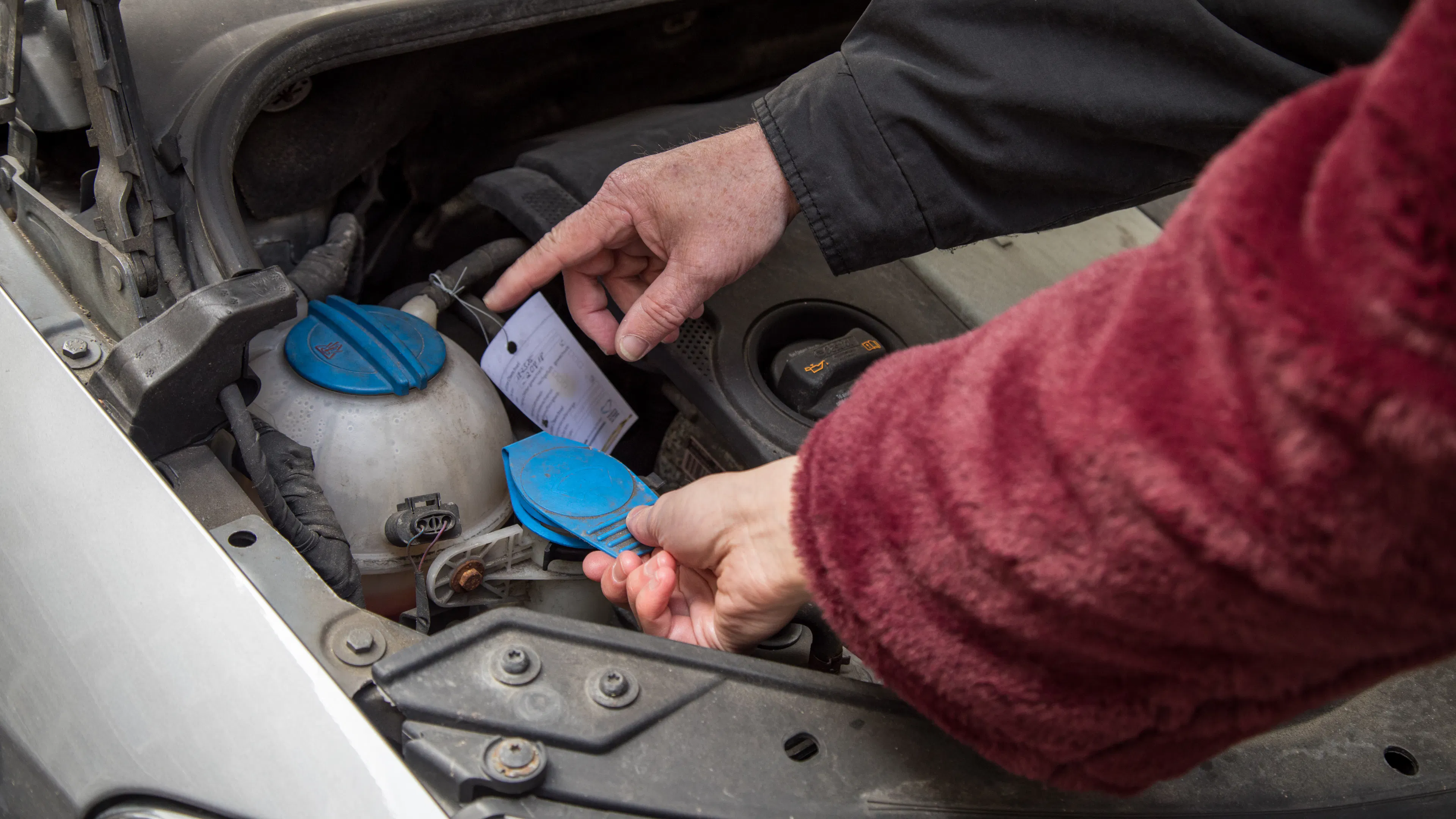 Zwei Personen sehen den Motorraum eines Autos an. Die eine Person zeigt auf den Kühlmittelbehälter, die andere fasst den Deckel des Scheibenwischwasserbehälters an.
