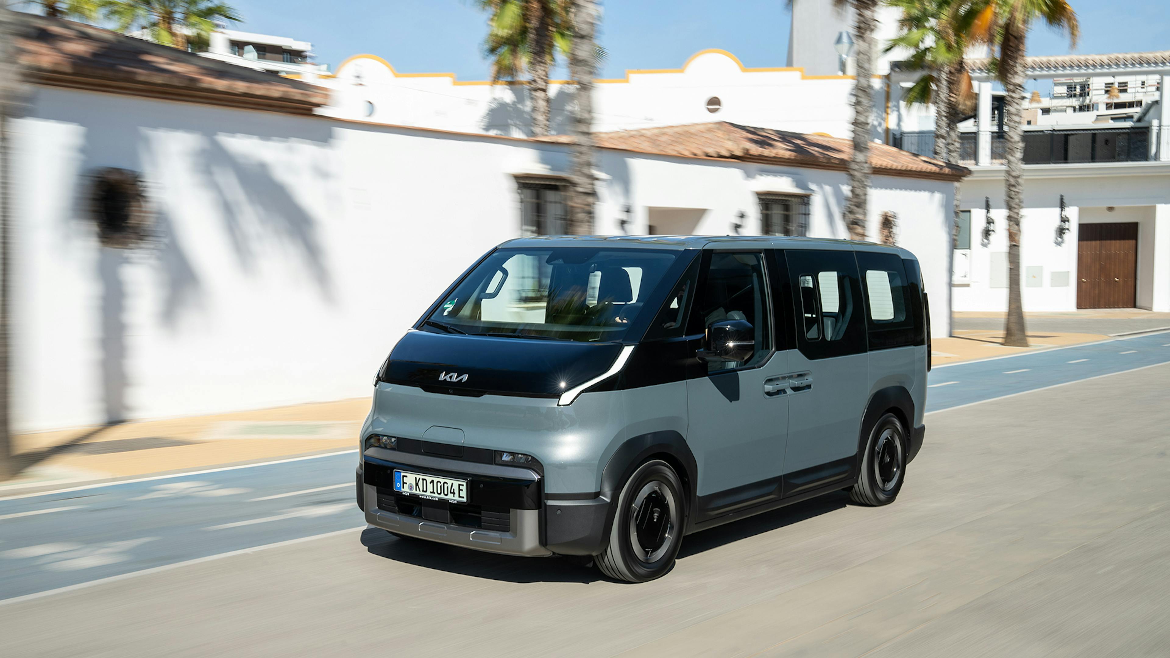 Ein blaugrauer Kia PV5 Passenger fährt an einem Haus vorbei.