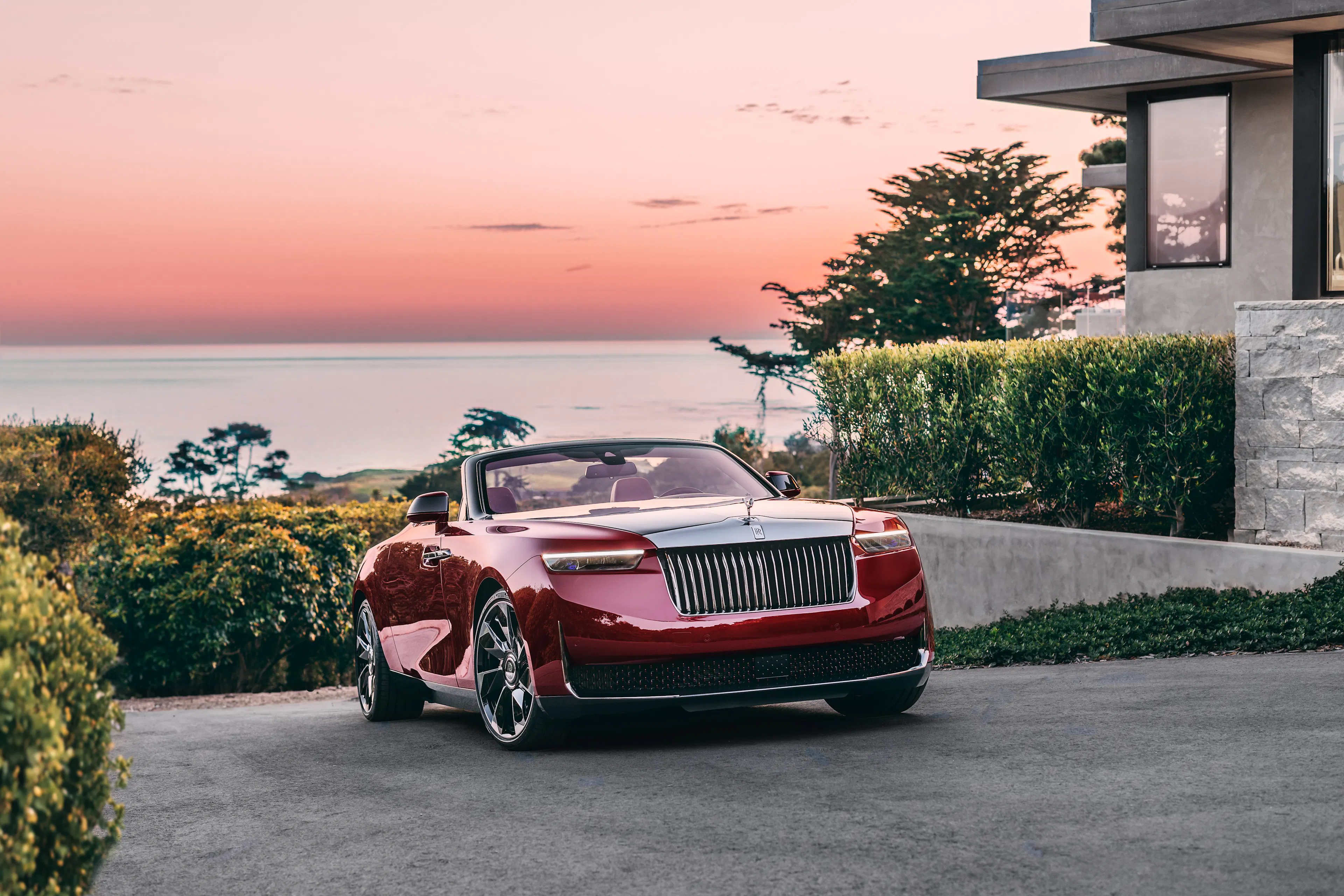 Ein Rolls-Royce La Rose Noire Droptail steht vor einer Villa an der Küste, im Hintergrund ist das Meer zu sehen.