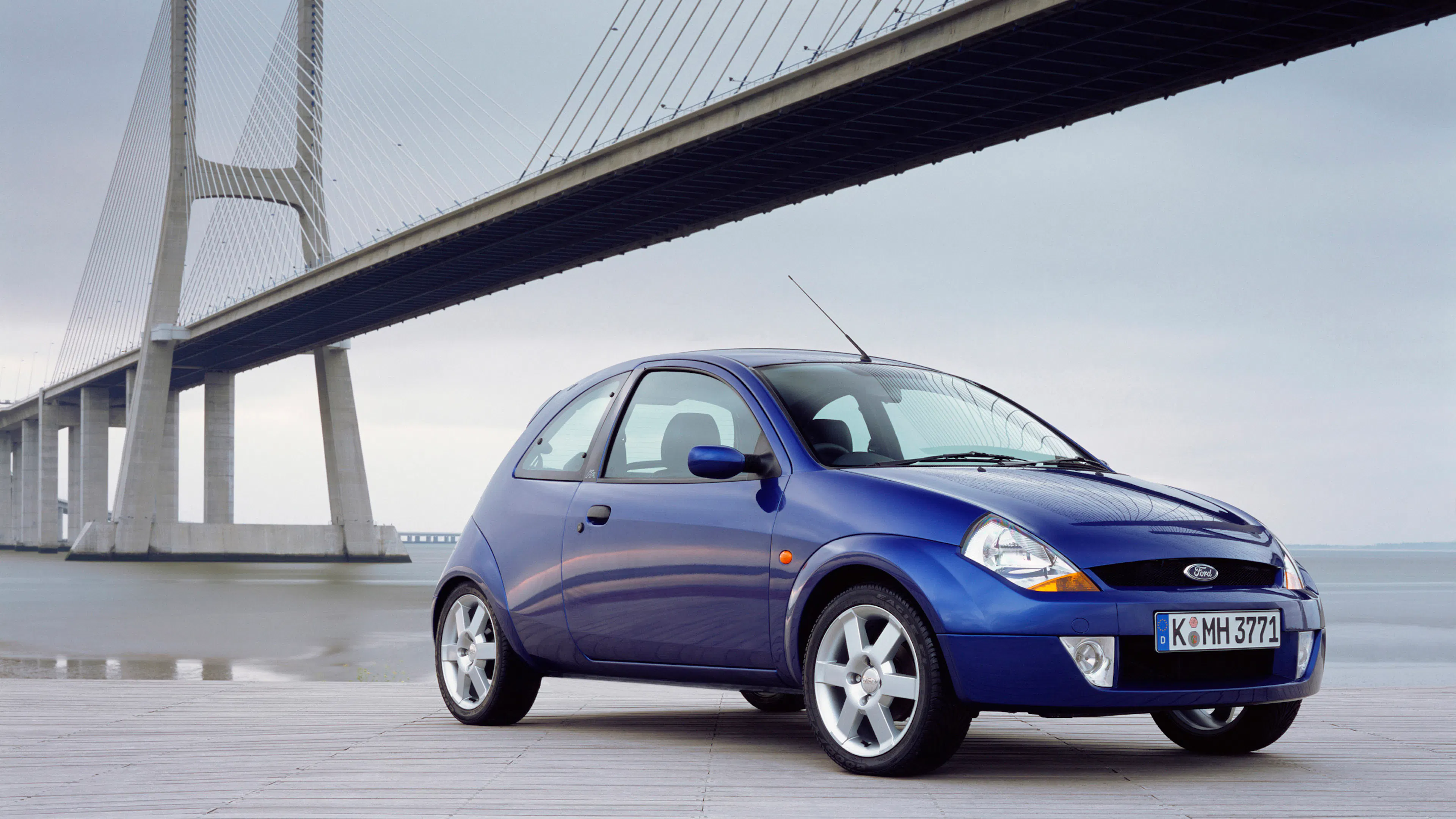 Ein blauer Ford Ka steht vor einer modernen Brücke.