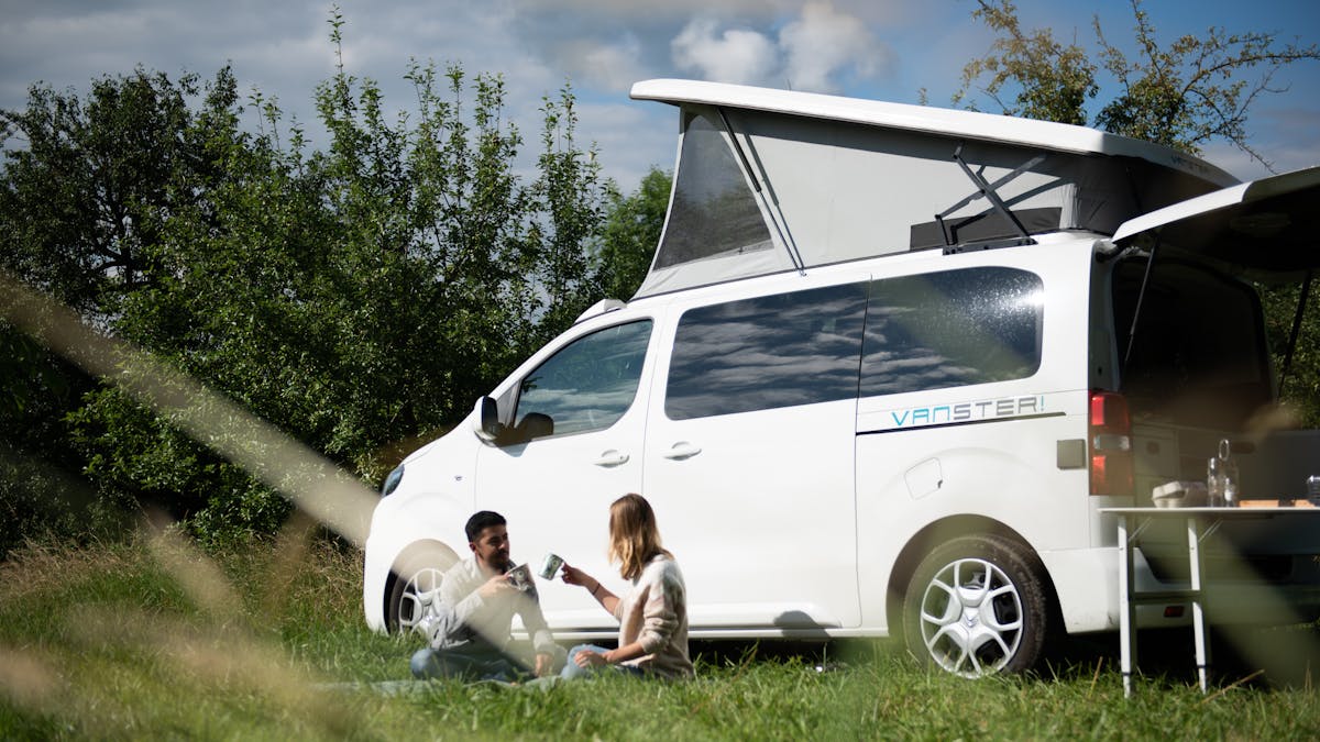 Zwei Personen sitzen vor einem weißen Pössl Vanster Campingbus im Gras