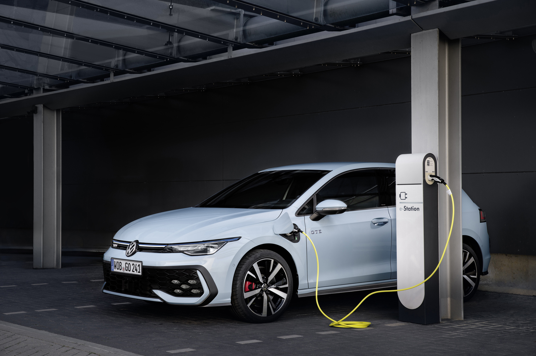Hellblauer VW Golf GTE in der Schrägansicht, angeschlossen an eine weiße e-Station-Ladesäule in einem Parkhaus, gelbes Ladekabel.