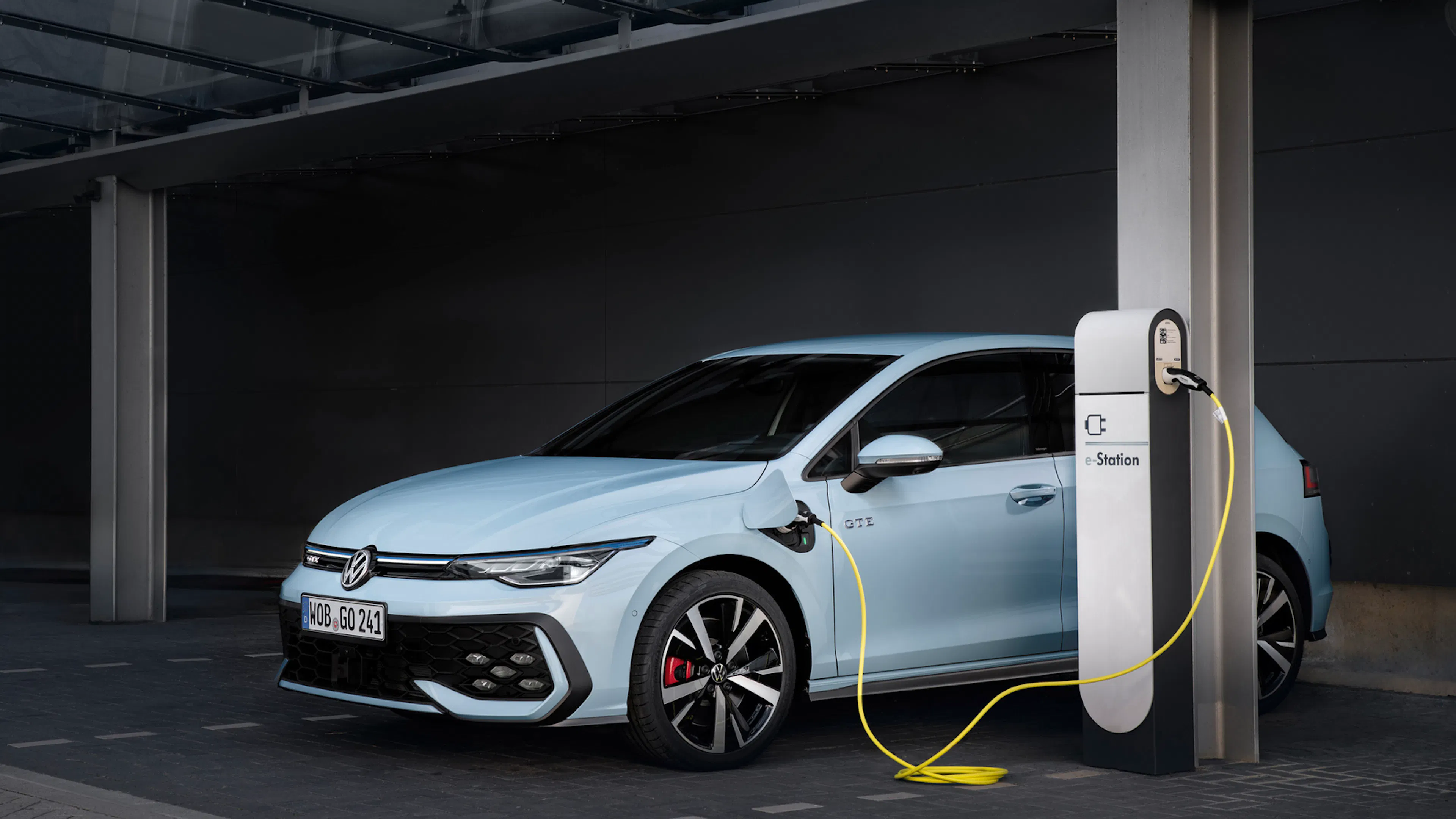 Hellblauer VW Golf GTE in der Schrägansicht, angeschlossen an eine weiße e-Station-Ladesäule in einem Parkhaus, gelbes Ladekabel.