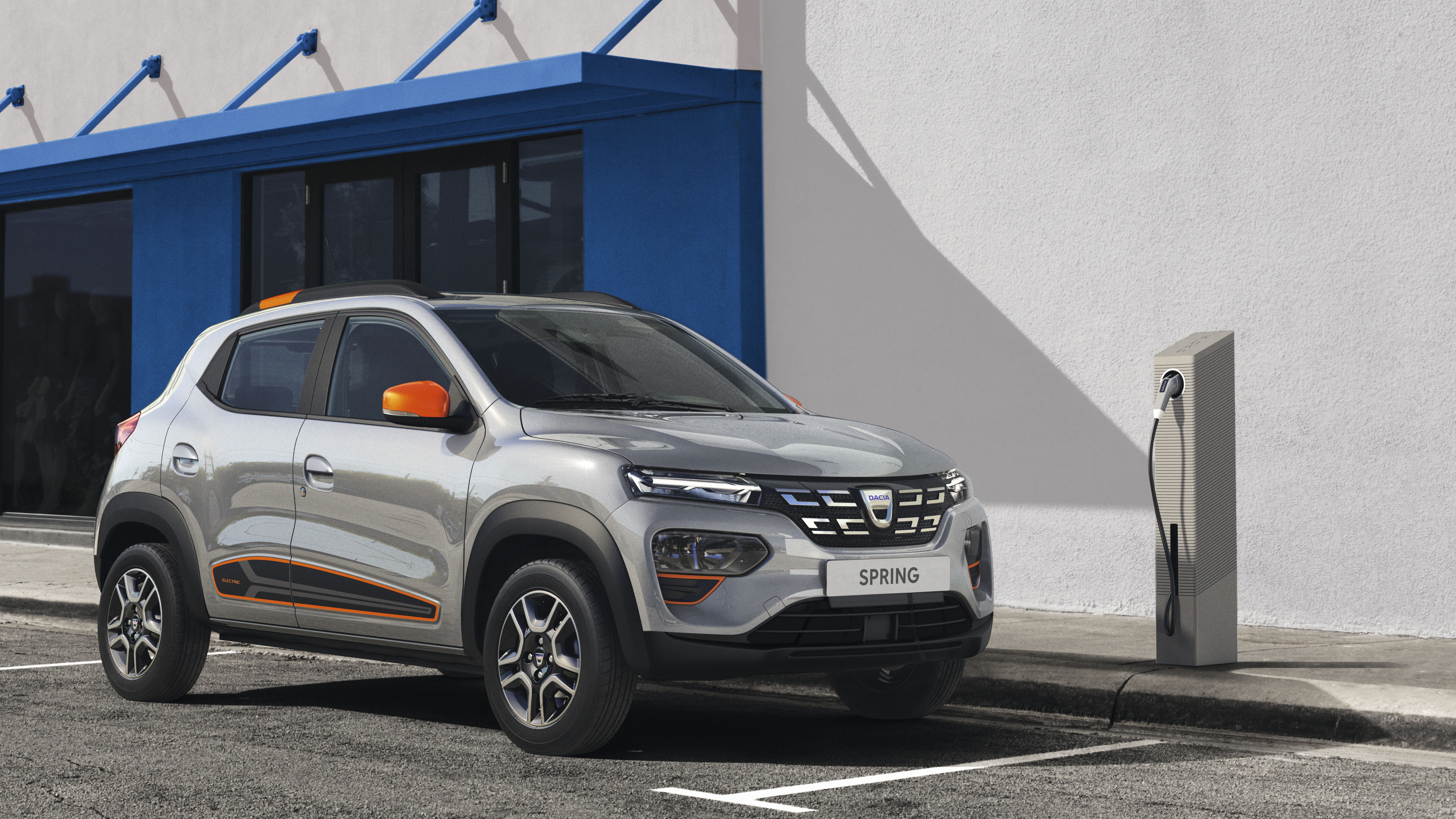 Ein silberner Dacia Spring Electric mit orangefarbenen Zierelementen steht an einer Ladesäule.