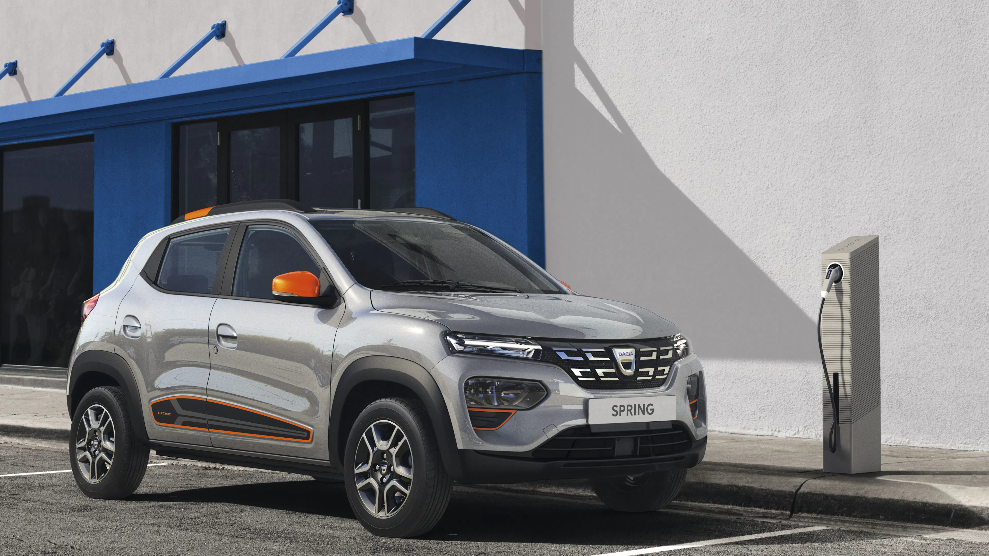 Ein silberner Dacia Spring Electric mit orangefarbenen Zierelementen steht an einer Ladesäule.