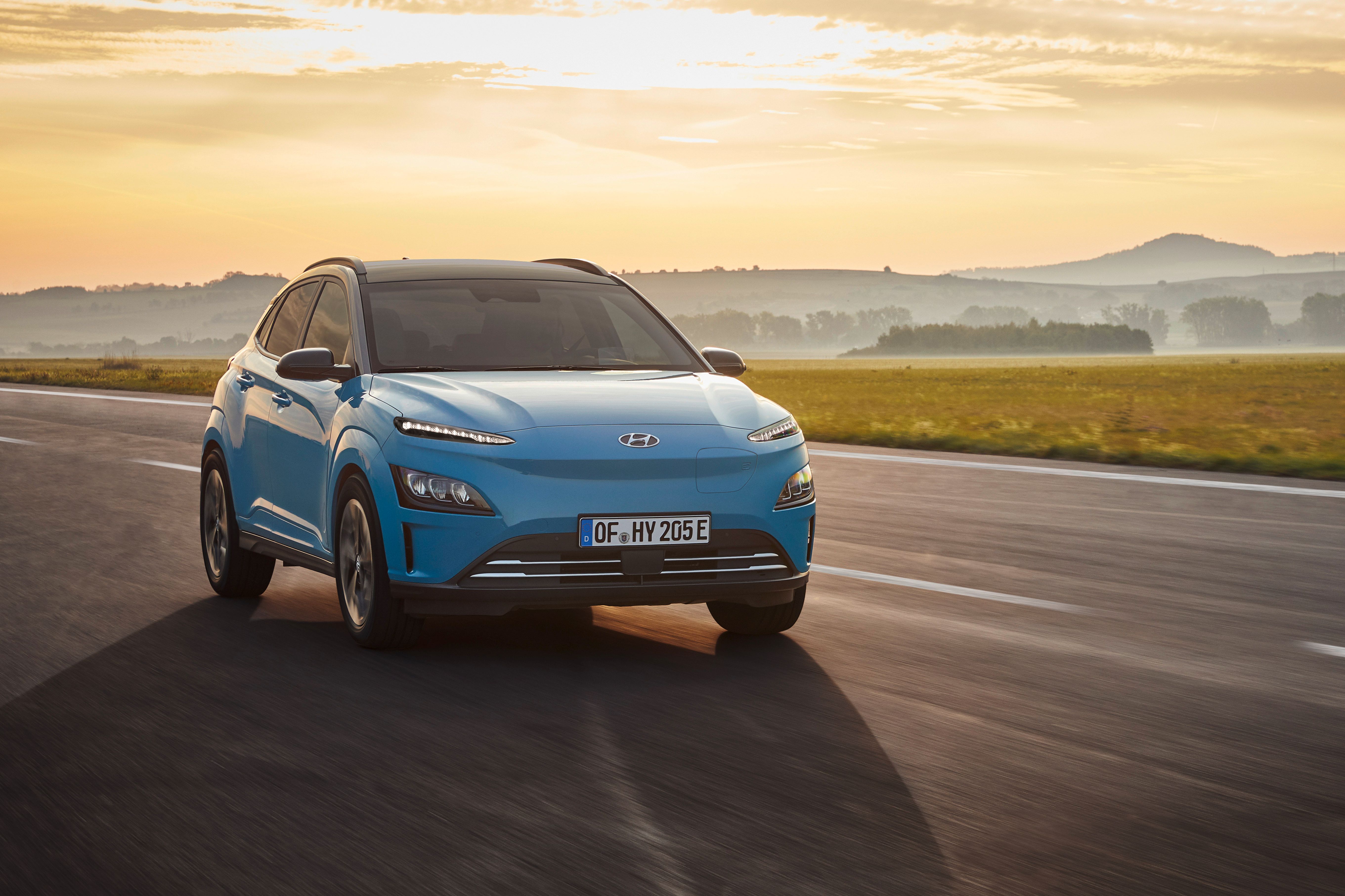 Ein blauer Hyundai Kona Elektro fährt auf einer Straße bei Sonnenuntergang.