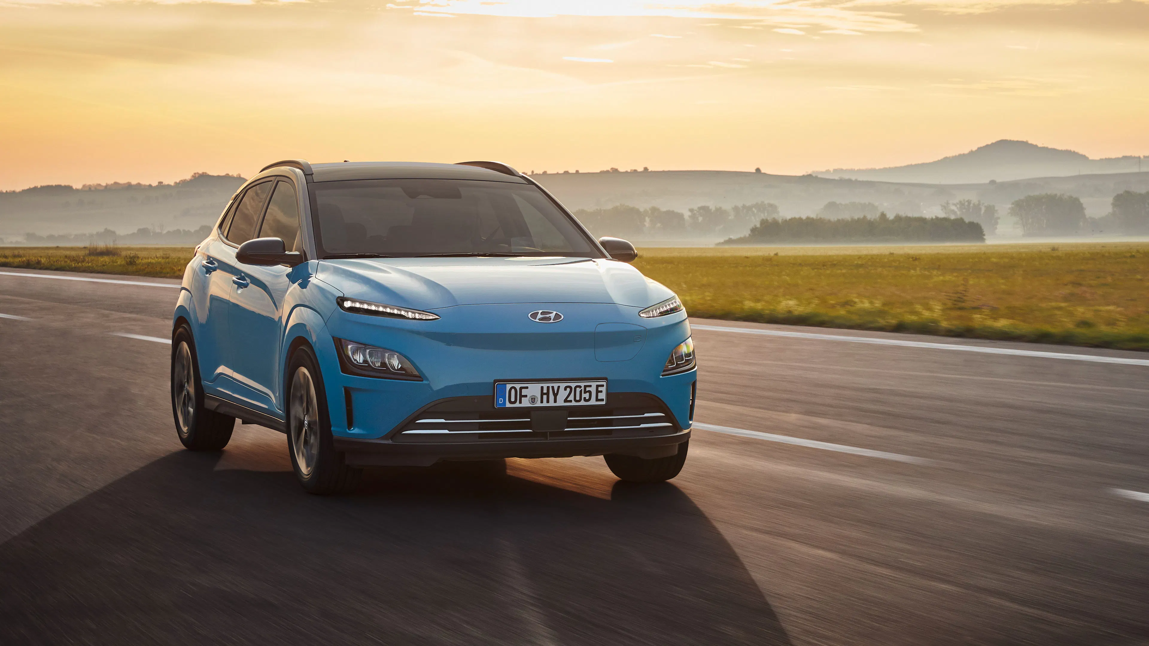 Ein blauer Hyundai Kona Elektro fährt auf einer Straße bei Sonnenuntergang.