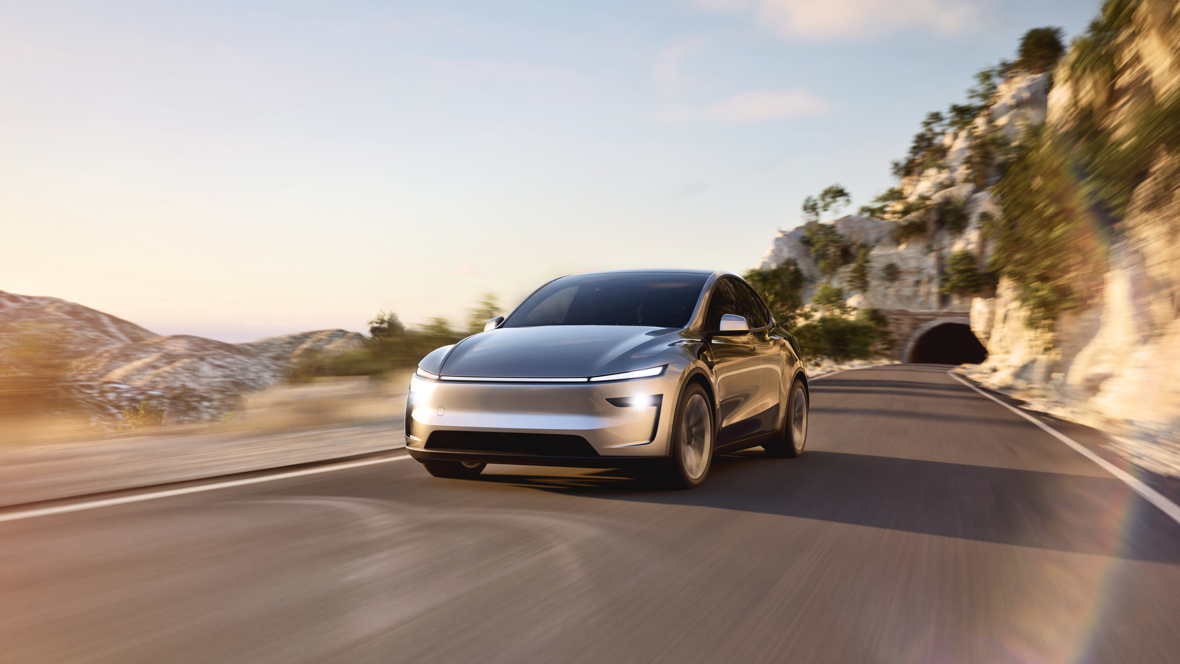 Ein silberner Tesla Model Y fährt auf einer sonnigen Bergstraße.