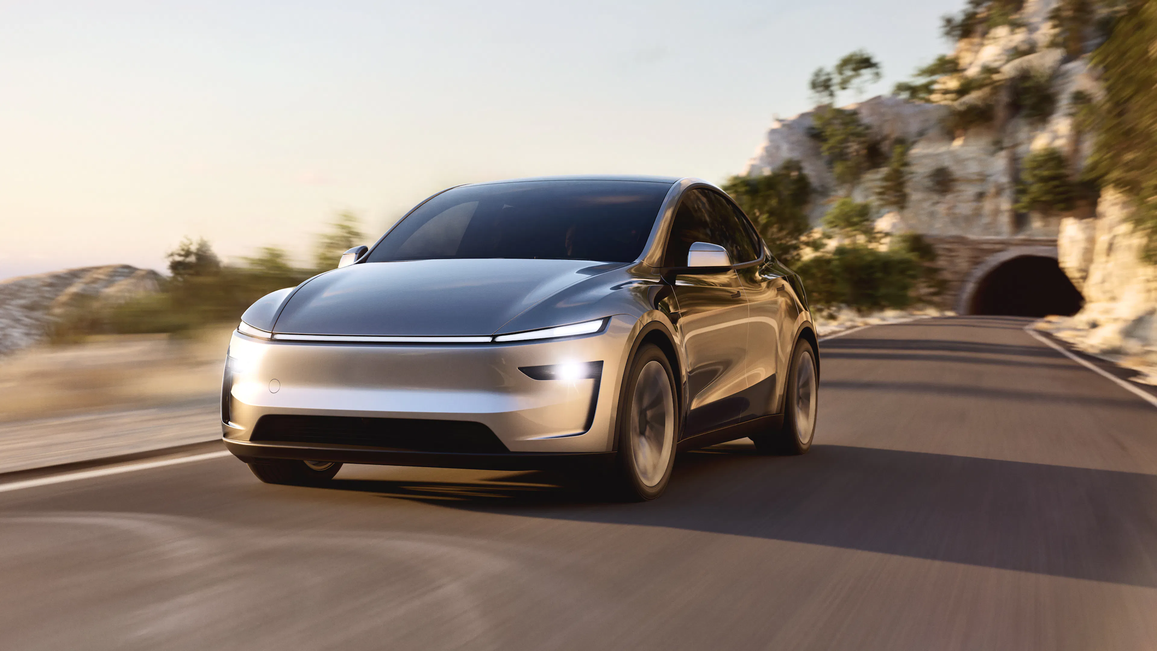 Ein silberner Tesla Model Y fährt auf einer sonnigen Bergstraße.