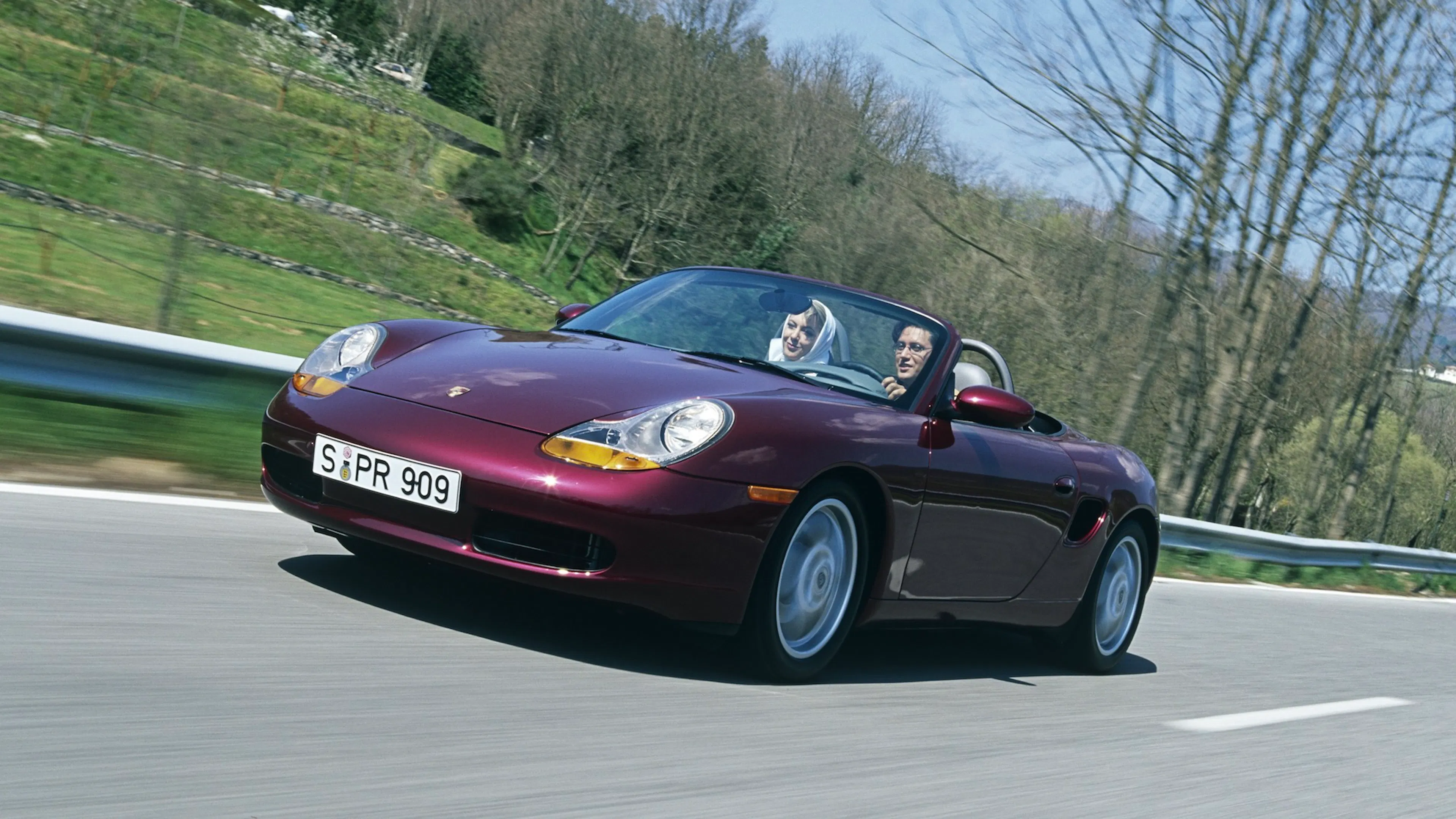 Ein weinroter Porsche Boxster 986 fährt auf einer Landstraße.