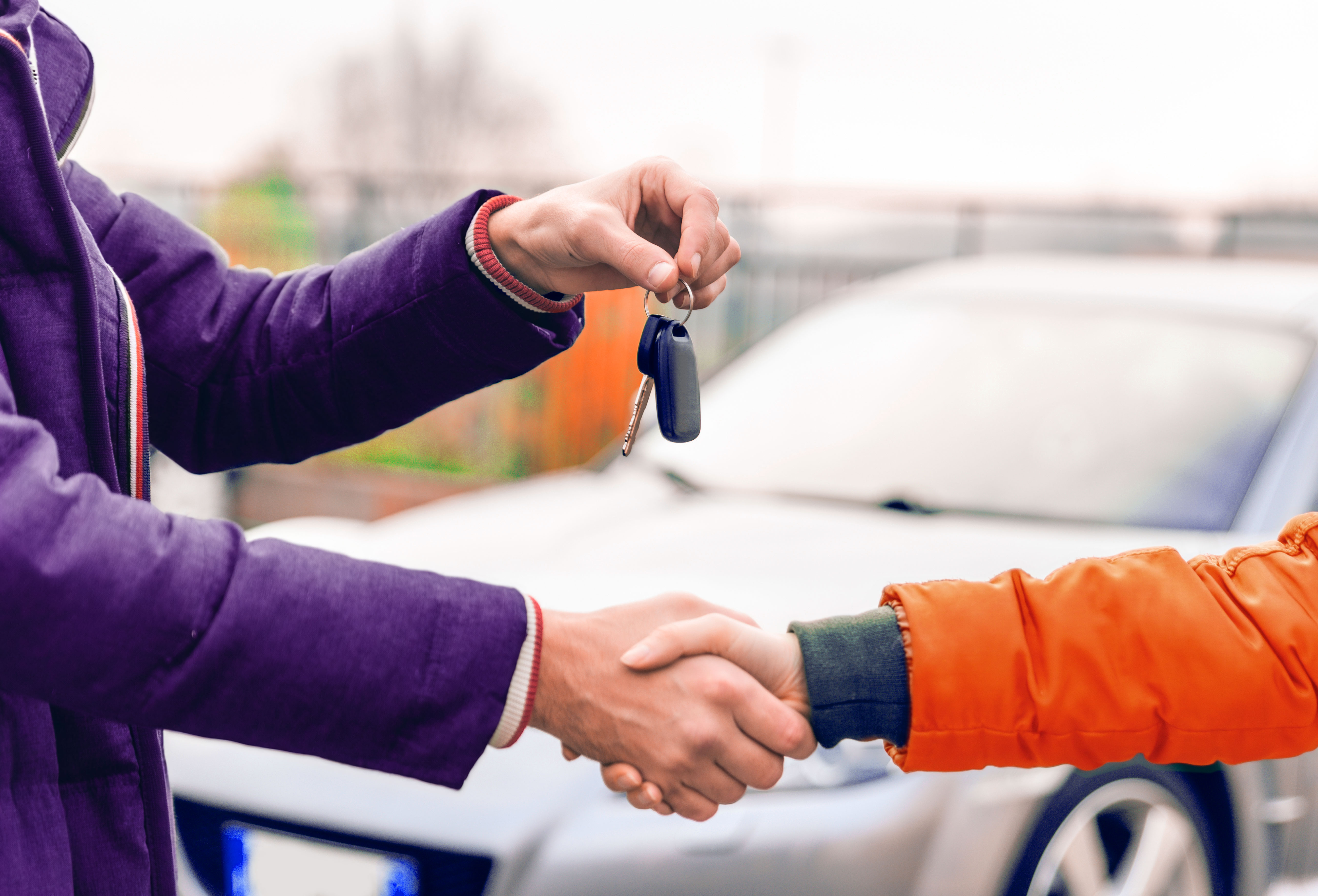 Zwei Personen reichen sich vor einem Fahrzeug die Hand. Eine übergibt der anderen einen Autoschlüssel.