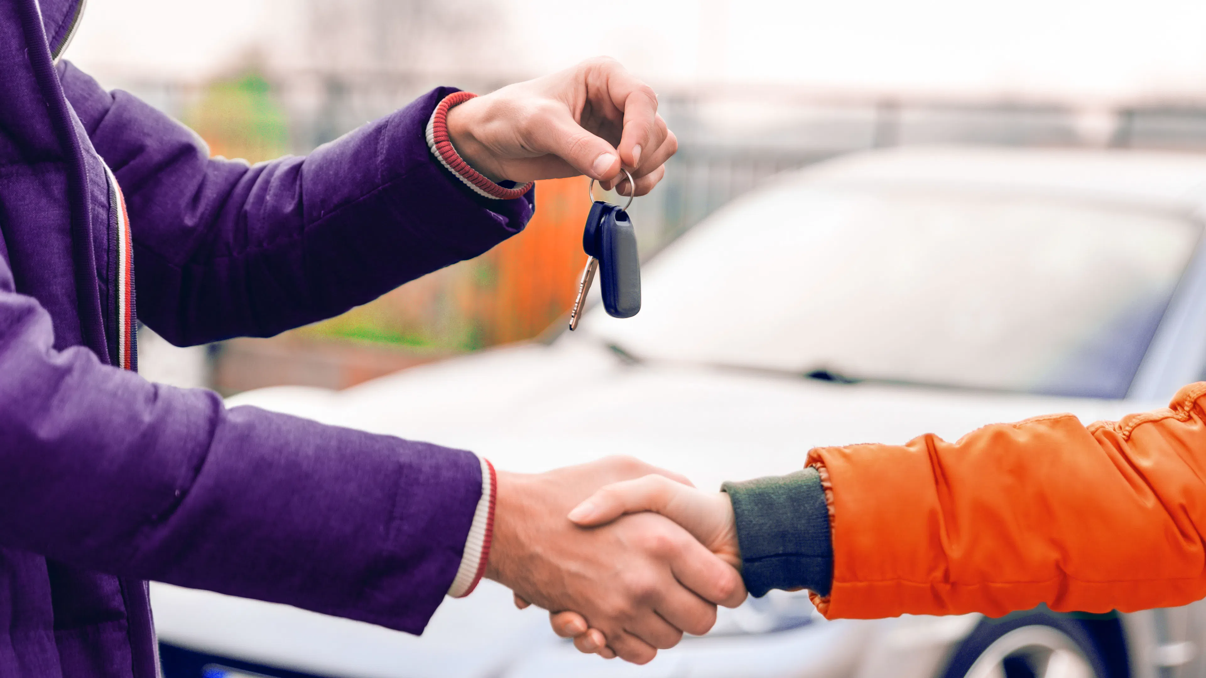 Zwei Personen reichen sich vor einem Fahrzeug die Hand. Eine übergibt der anderen einen Autoschlüssel.