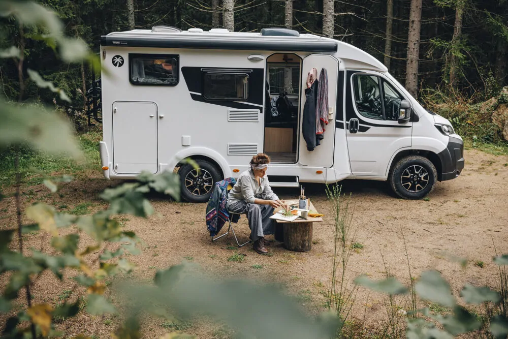 Eine Frau sitzt auf einem Campingstuhl vor einem weißen Sunlight-V60-Wohnmobil, das auf einem Waldparkplatz steht, Schiebetür geöffnet.