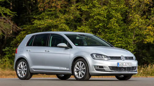 Ein silberner VW Golf steht auf einer Straße vor Bäumen.