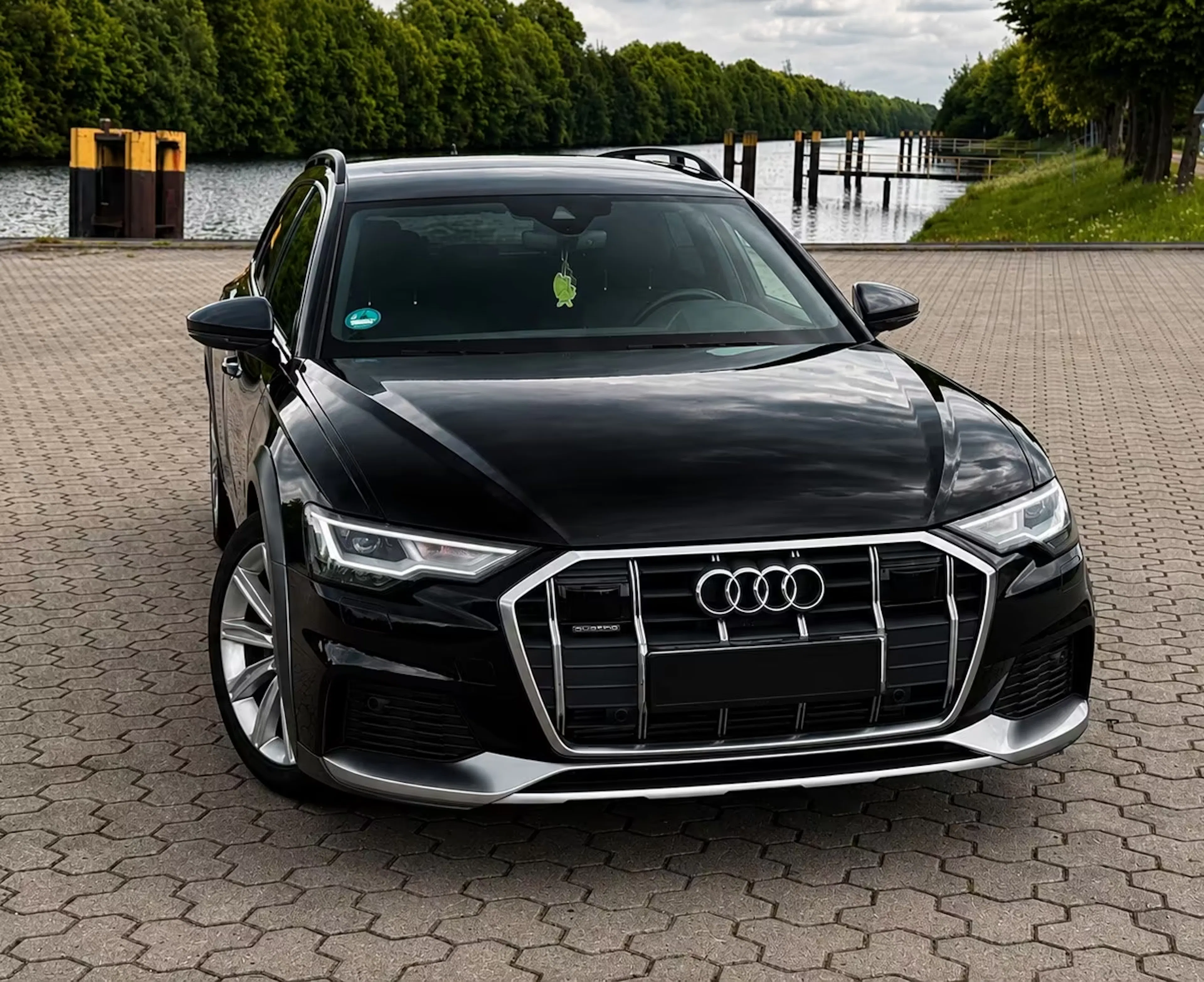 Ein schwarzer Audi A6 Allroad steht auf einem gepflastertem Platz vor einem See.