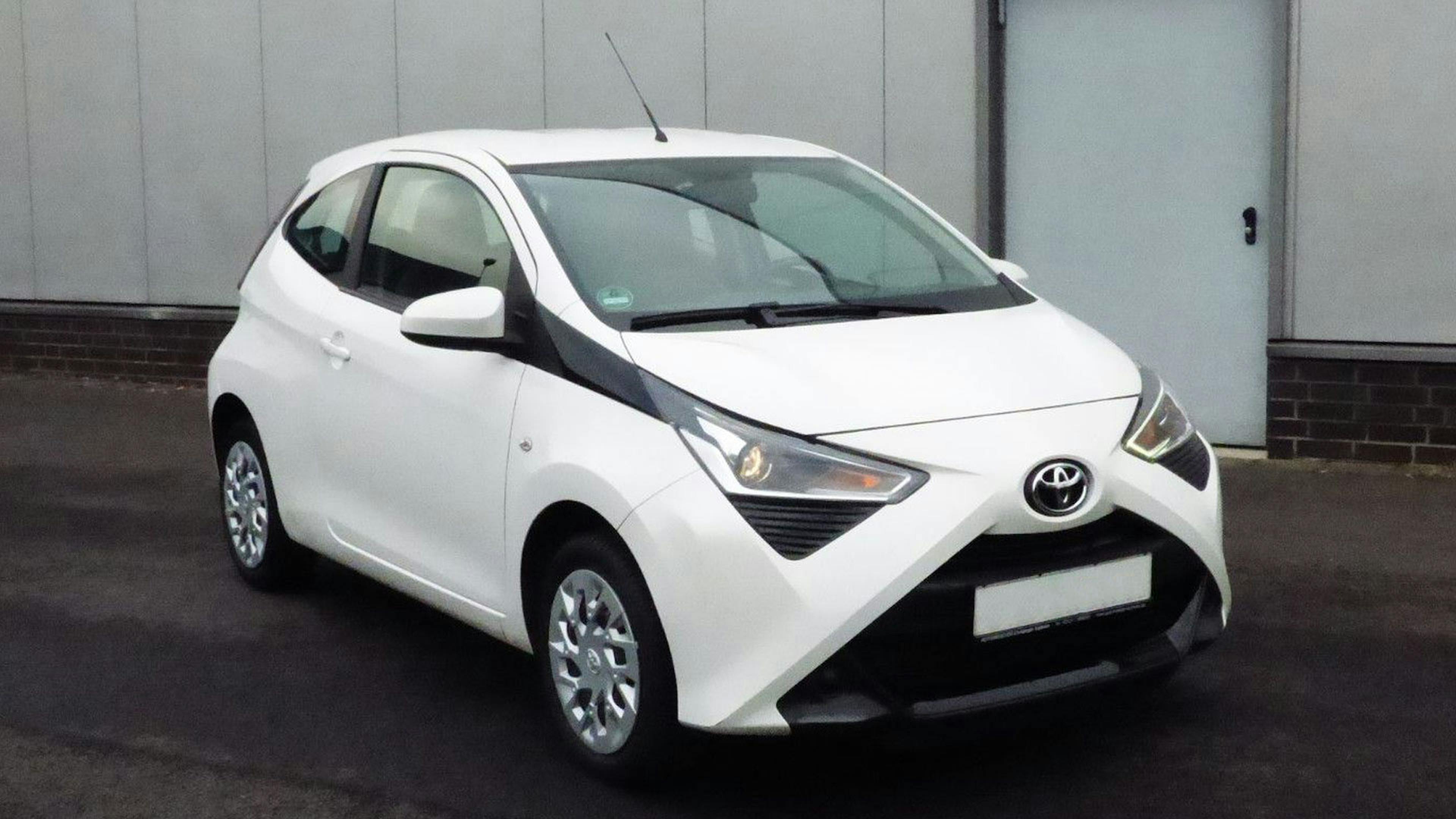 Ein weißer Toyota Aygo Kleinstwagen steht vor einem grauen Gebäude.