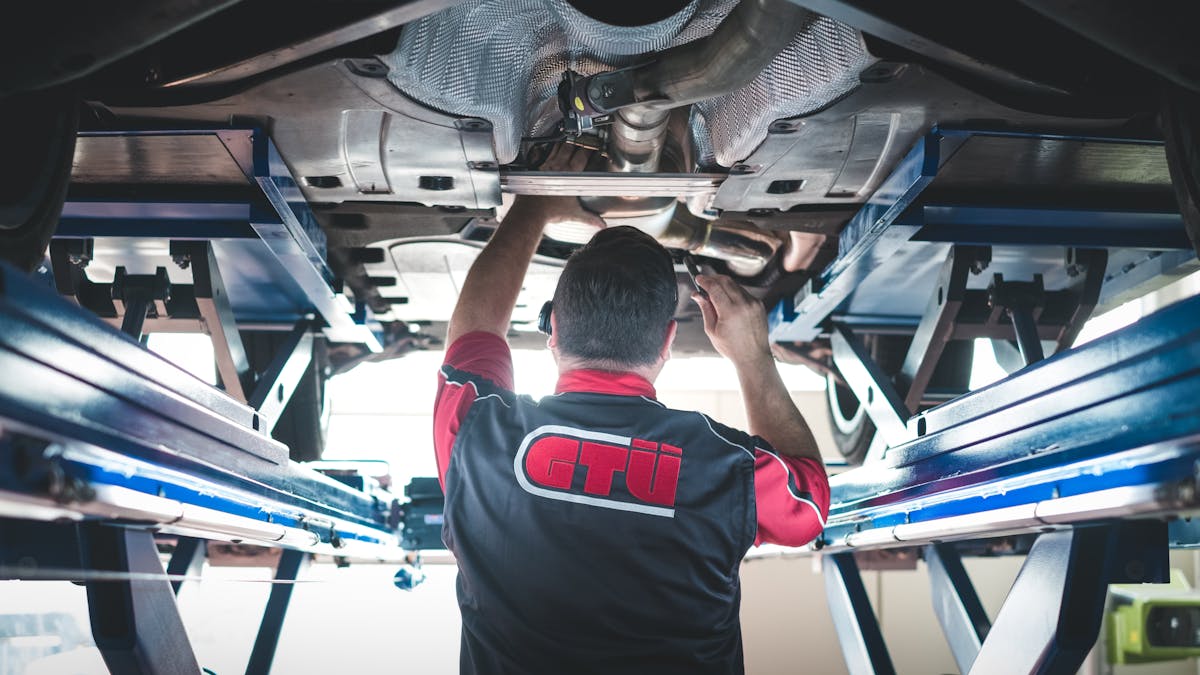 Ein Techniker der GTÜ steht unter einem Pkw, der auf einer Hebebühne steht, und führt eine Hauptuntersuchung durch