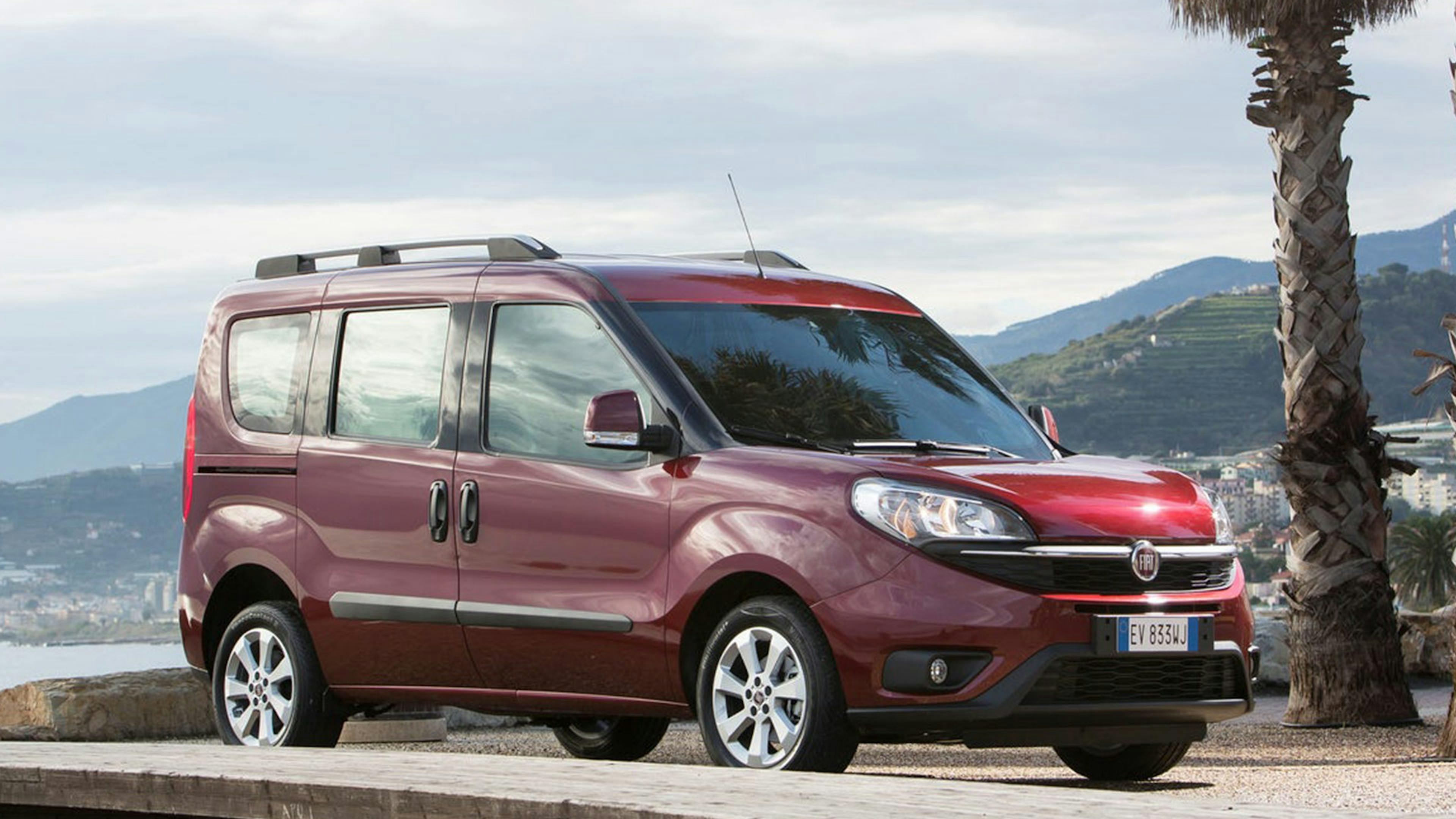 Ein dunkelroter Fiat Doblo Hochdachkombi fährt an einem grauen Gebäude vorbei.