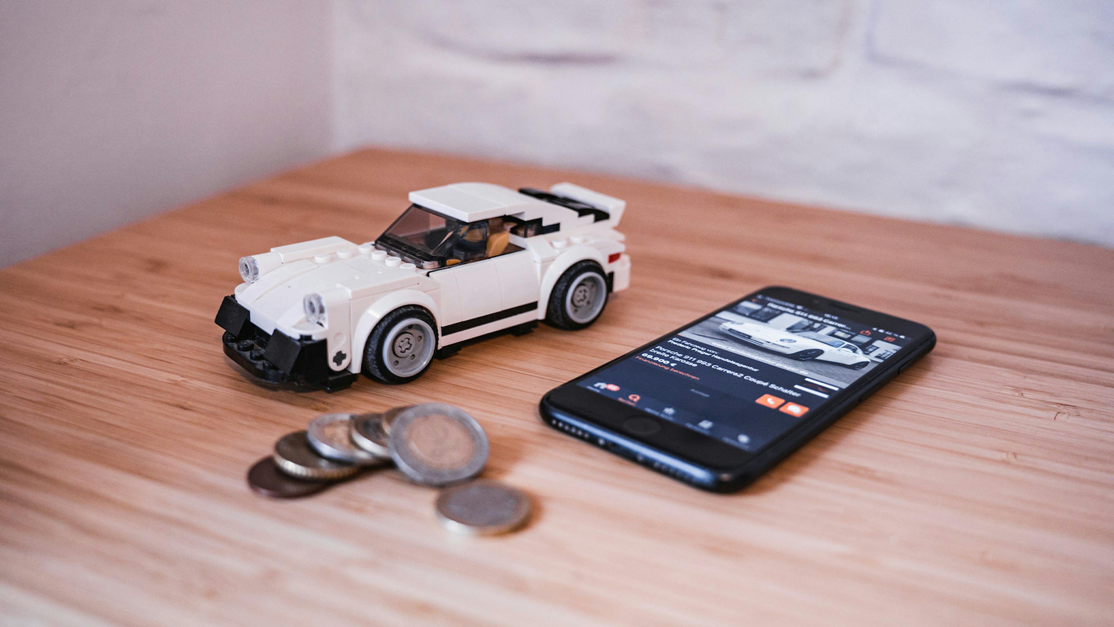 Stillleben mit einem Smartphone, einem weißen Spielzeugauto und diversen Geldmünzen