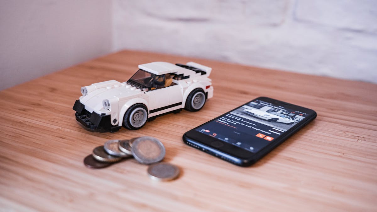 Stillleben mit einem Smartphone, einem weißen Spielzeugauto und diversen Geldmünzen
