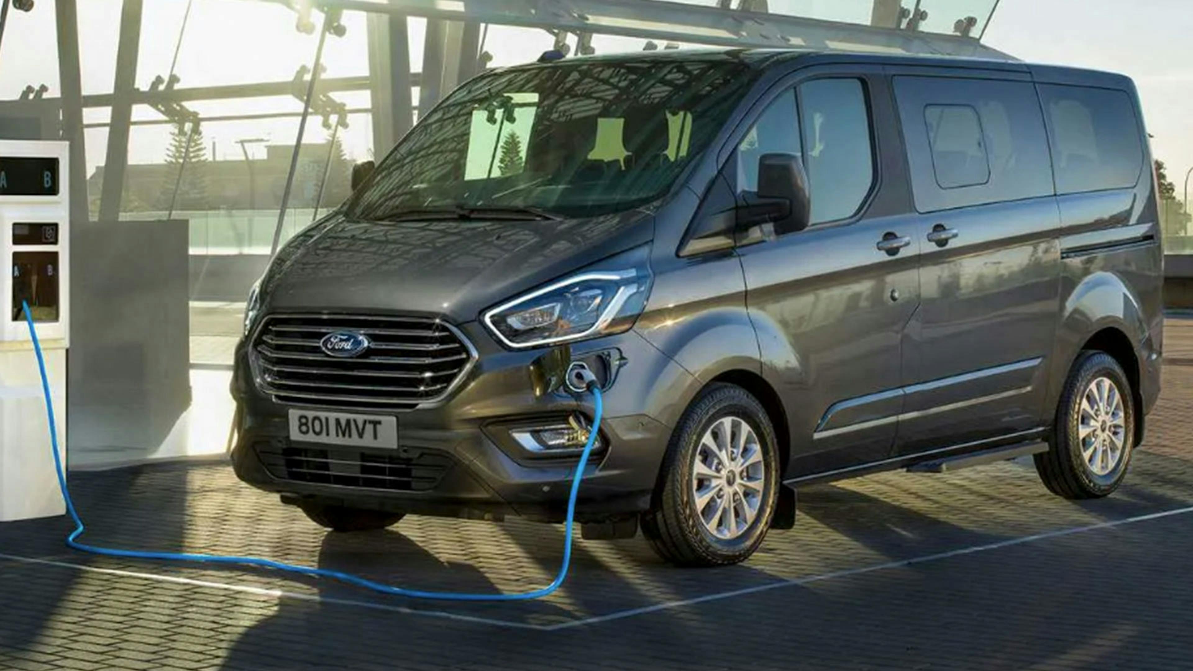 Ein grauer Ford Tourneo Custom PHEV mit Hybrid-Antrieb wird an einer Ladesäule aufgeladen.