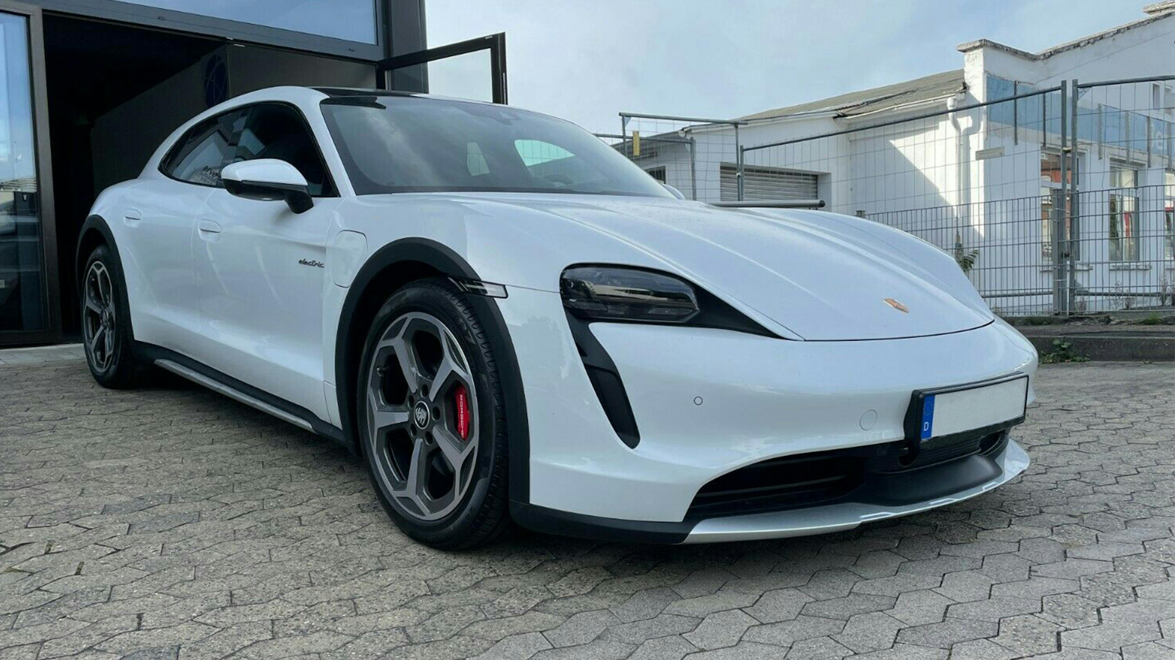 Ein weißer Porsche Taycan Cross Tourismo steht auf einem Parkplatz vor einer Halle