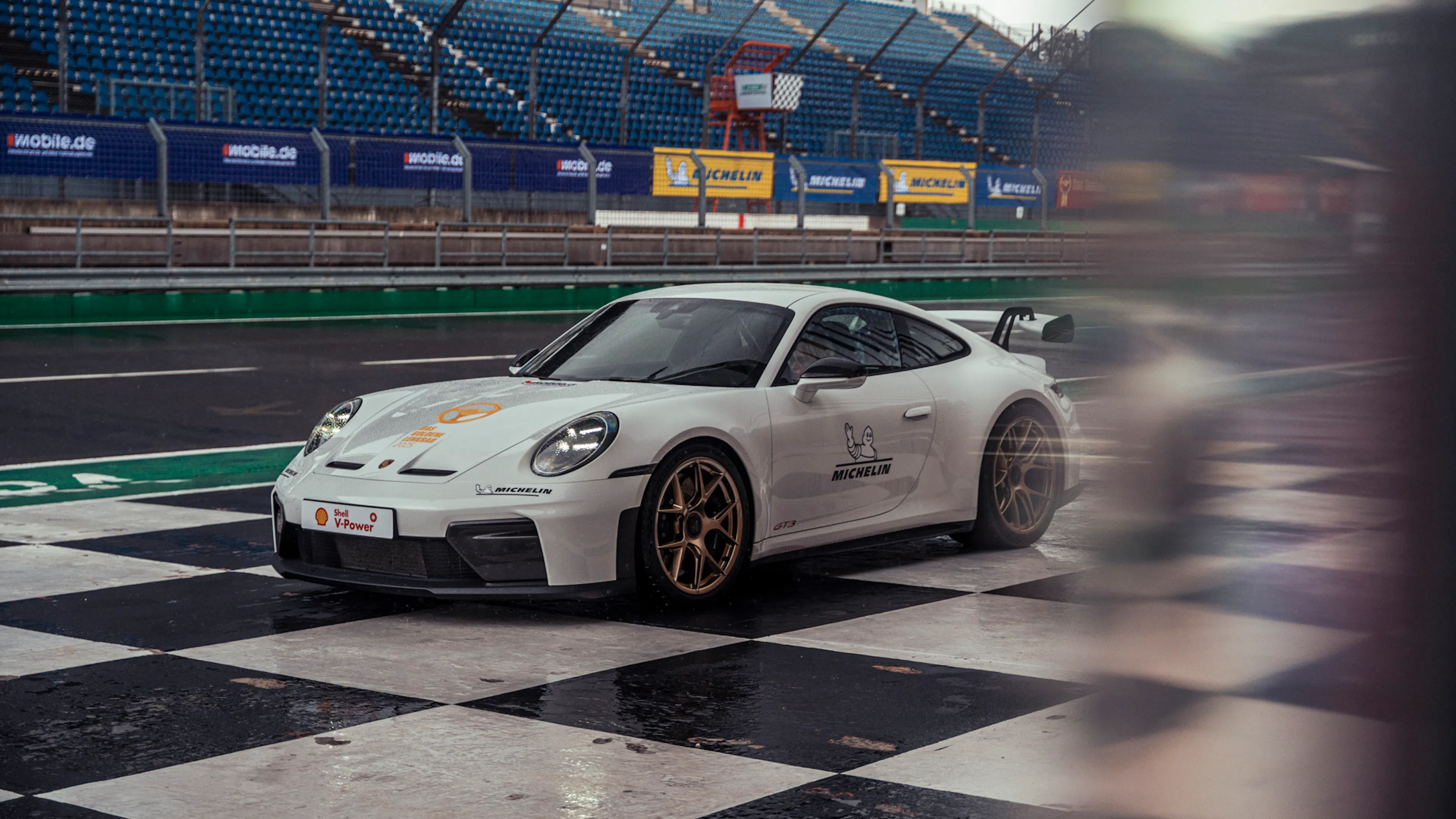 Weitwinkelaufnahme eines weißen Porsche 911 GT3 auf einem schwarz-weißen Schachbrettmuster vor dem Gebäude des DEKRA Lausitzrings.