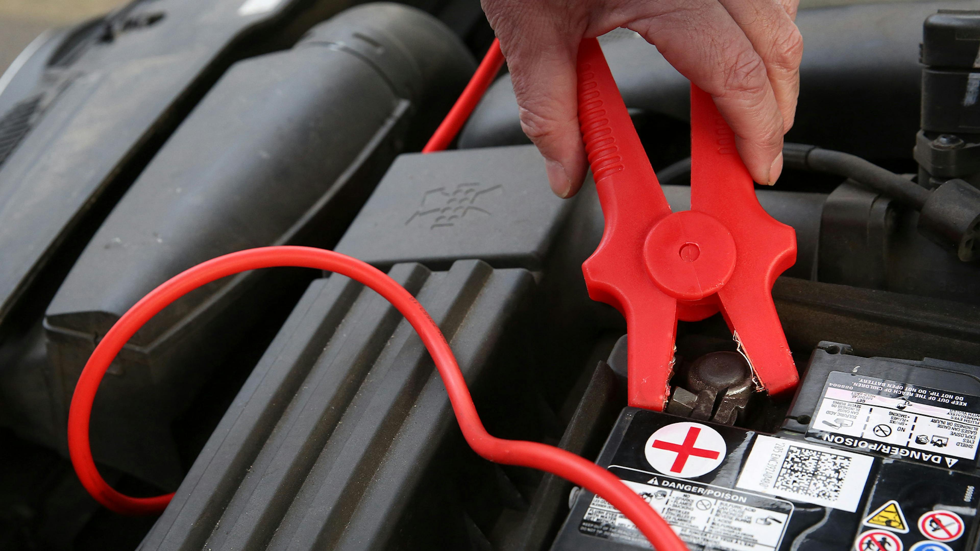 Eine Person befestigt die rote Klemme eines Starterkabels am Pluspol einer Autobatterie, um Starthilfe zu geben.